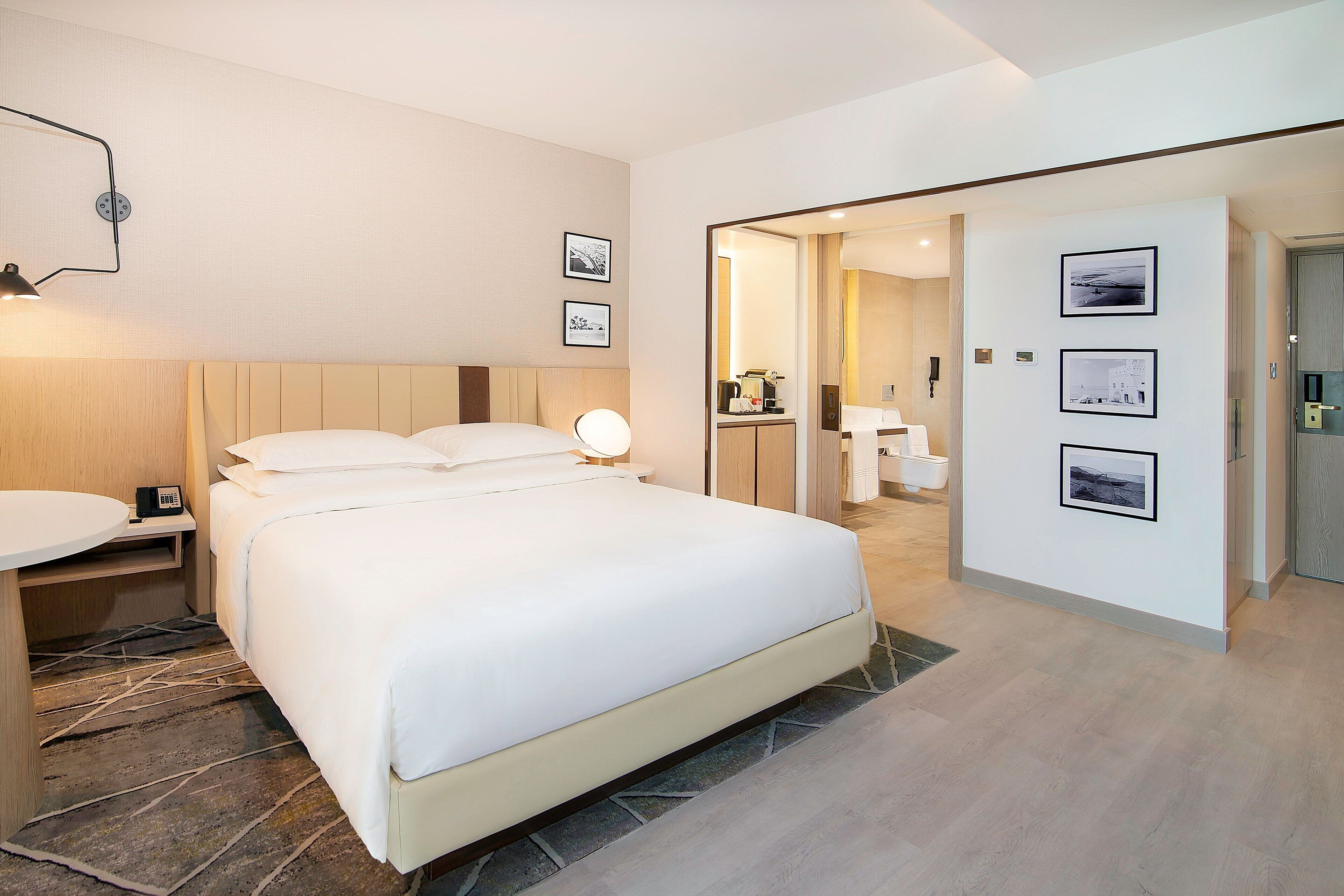 A modern hotel room with a large bed, bedside tables, a desk, and a mirror. There are framed pictures on the wall and a bathroom visible through an open door.
