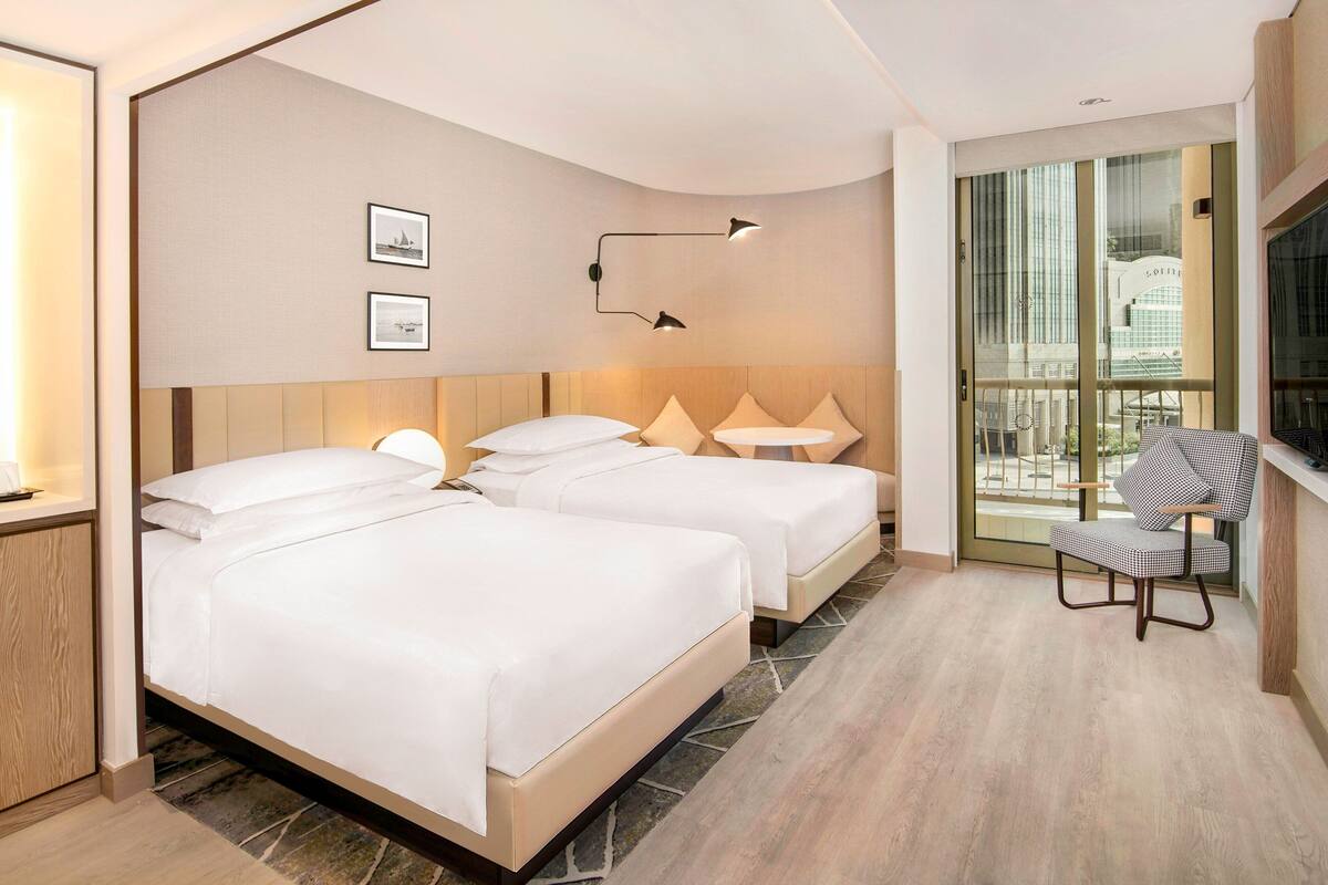 Habitación de hotel con dos camas, una mesita, una silla, un televisor y un ventanal con vista a la ciudad.