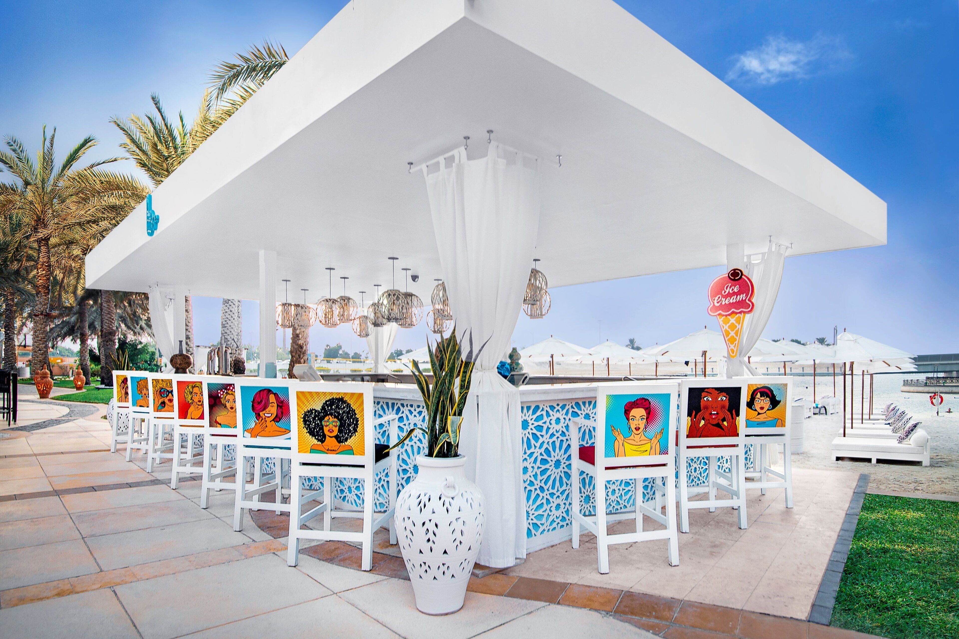 A covered outdoor area with a white canopy, a row of chairs with colorful artwork, and a large white vase with plants.
