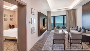 Habitación de hotel moderna con una cama grande, un sofá, dos sillones, una mesa de centro y un televisor. En la pared hay cuadros enmarcados y se ve el mar a través de la ventana.