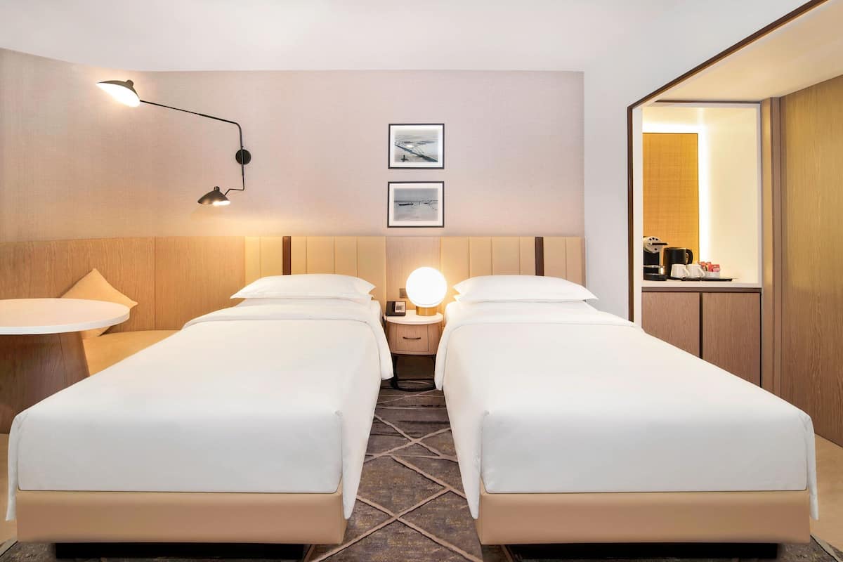 Una habitación de hotel con dos camas individuales, una mesa redonda, una mesita de noche con lámpara y un espejo pequeño y redondo.
