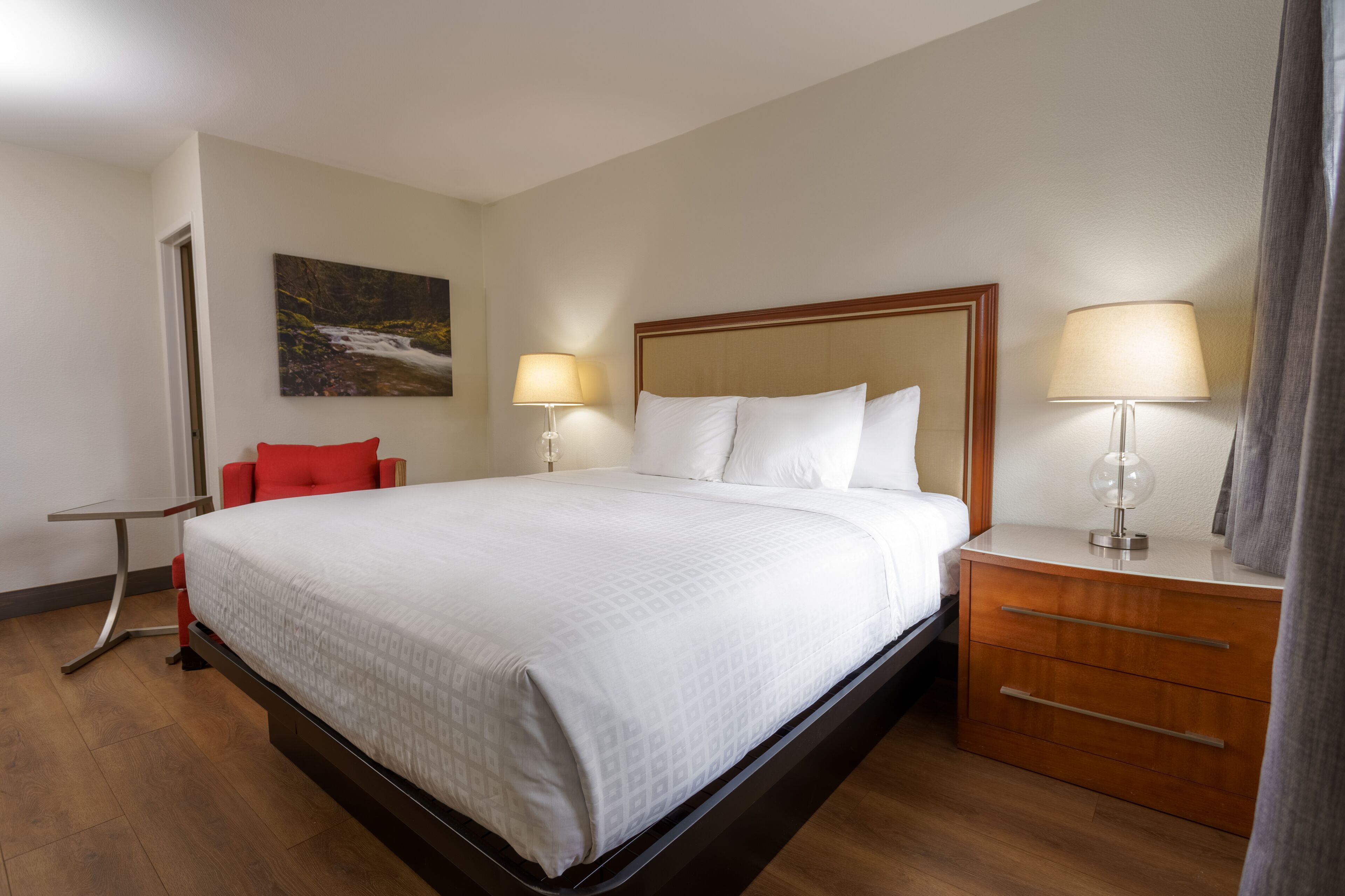 Una habitación de hotel con una cama, mesitas de noche, una silla roja y un cuadro en la pared.