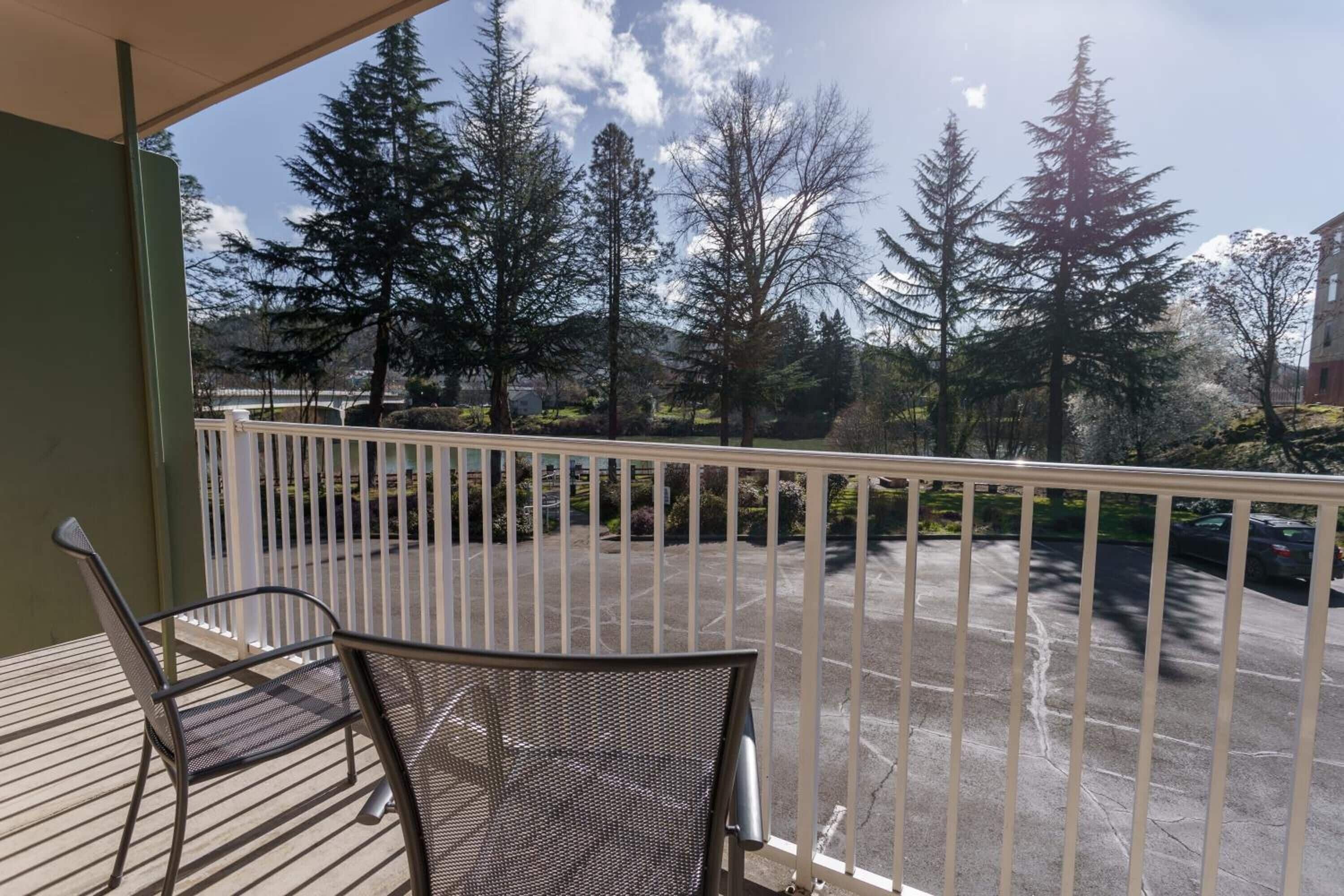 A balcony with two chairs, a view of a parking lot, and tall trees in the background.