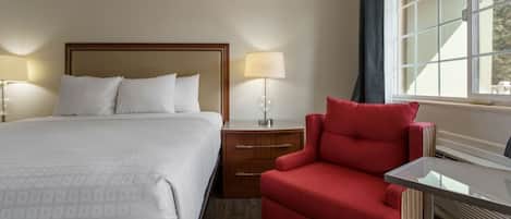 Habitación de hotel con una cama grande, un sillón rojo, una mesita de noche con lámpara y un ventanal con cortinas.