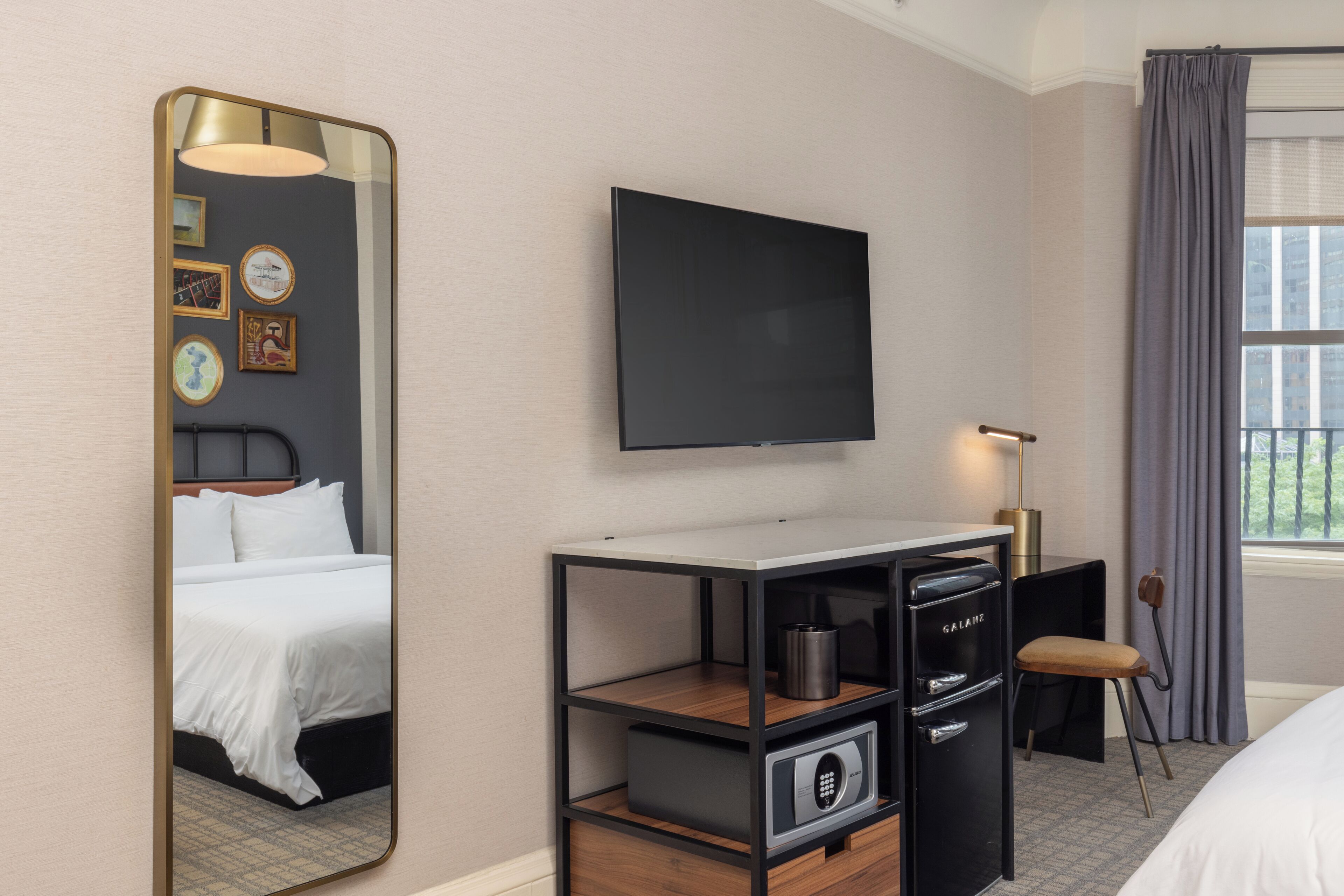 Ett hotellrum med en säng, en tv, ett skrivbord med en lampa, en stol och ett fönster med gardiner.