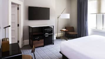 A hotel room with a bed, a desk with a computer, a TV, a chair, a lamp, and a window with curtains.