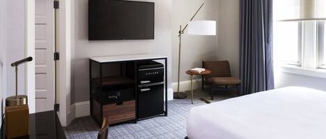 A hotel room with a bed, a desk with a computer, a TV, a chair, a lamp, and a window with curtains.