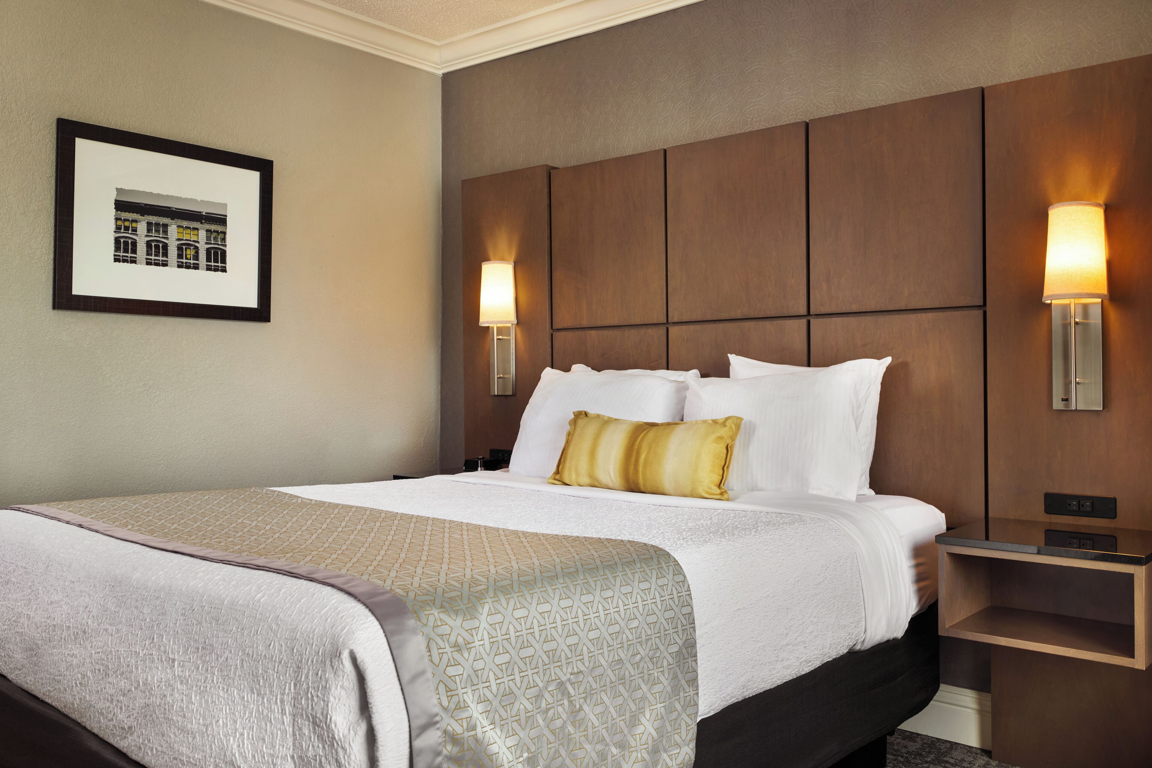 A hotel room with a large bed, a headboard with panels, two wall-mounted lamps, a framed picture on the wall, and a bedside table.
