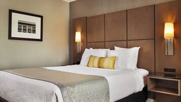 A hotel room with a large bed, a headboard with panels, two wall-mounted lamps, a framed picture on the wall, and a bedside table.