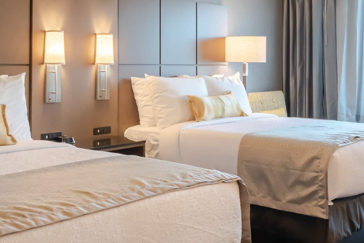 Quarto de hotel com duas camas, cada uma com roupa de cama branca e cabeceira. Há luminárias fixadas na parede, uma de cada lado das camas.