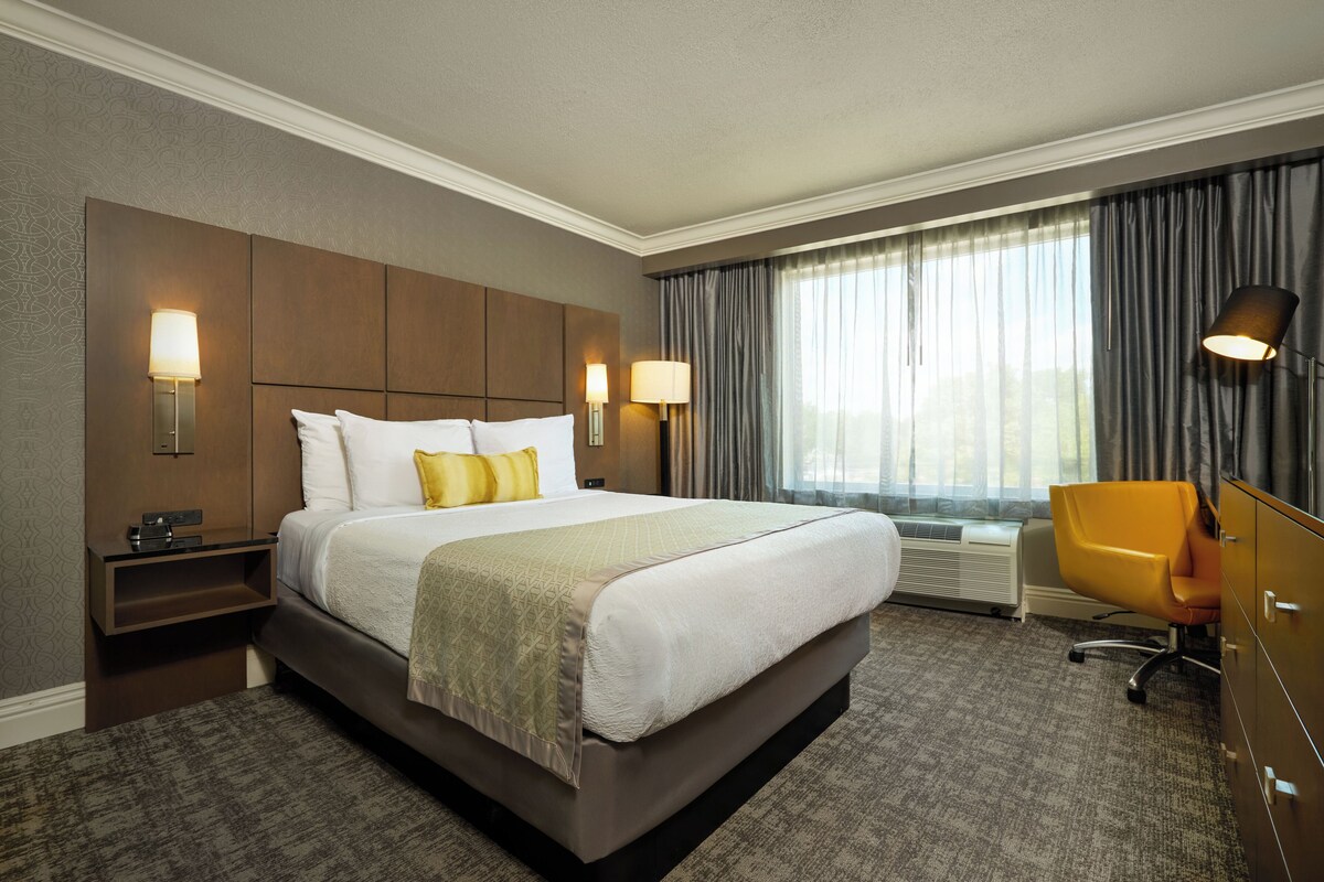Quarto de hotel com uma cama grande, duas luminárias de cabeceira, uma escrivaninha, uma cadeira e uma janela com cortinas.