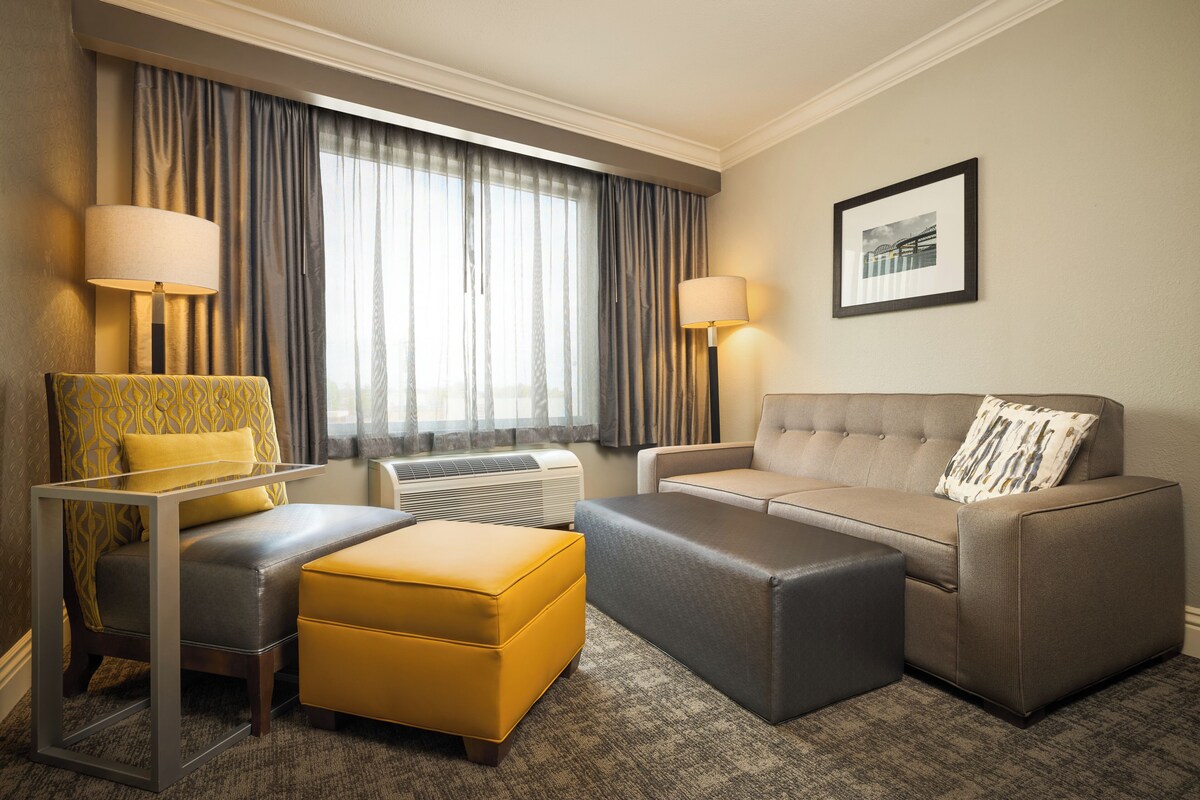 Quarto de hotel com sofá, banqueta, poltrona e mesa com luminárias.