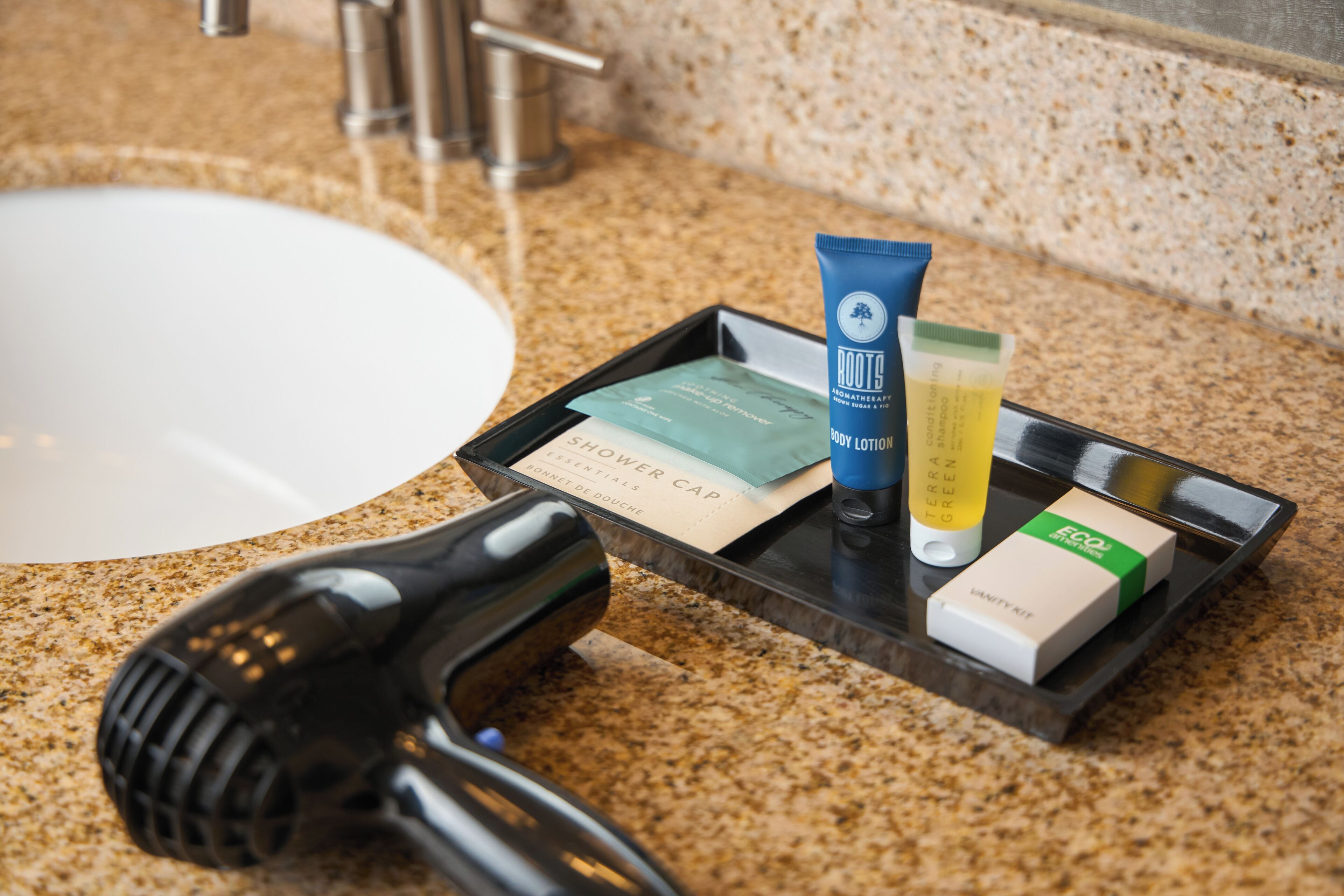 Una encimera de baño con un secador de pelo, una bandeja con artículos de aseo personal, incluyendo una loción corporal, un champú verde y un neceser.
