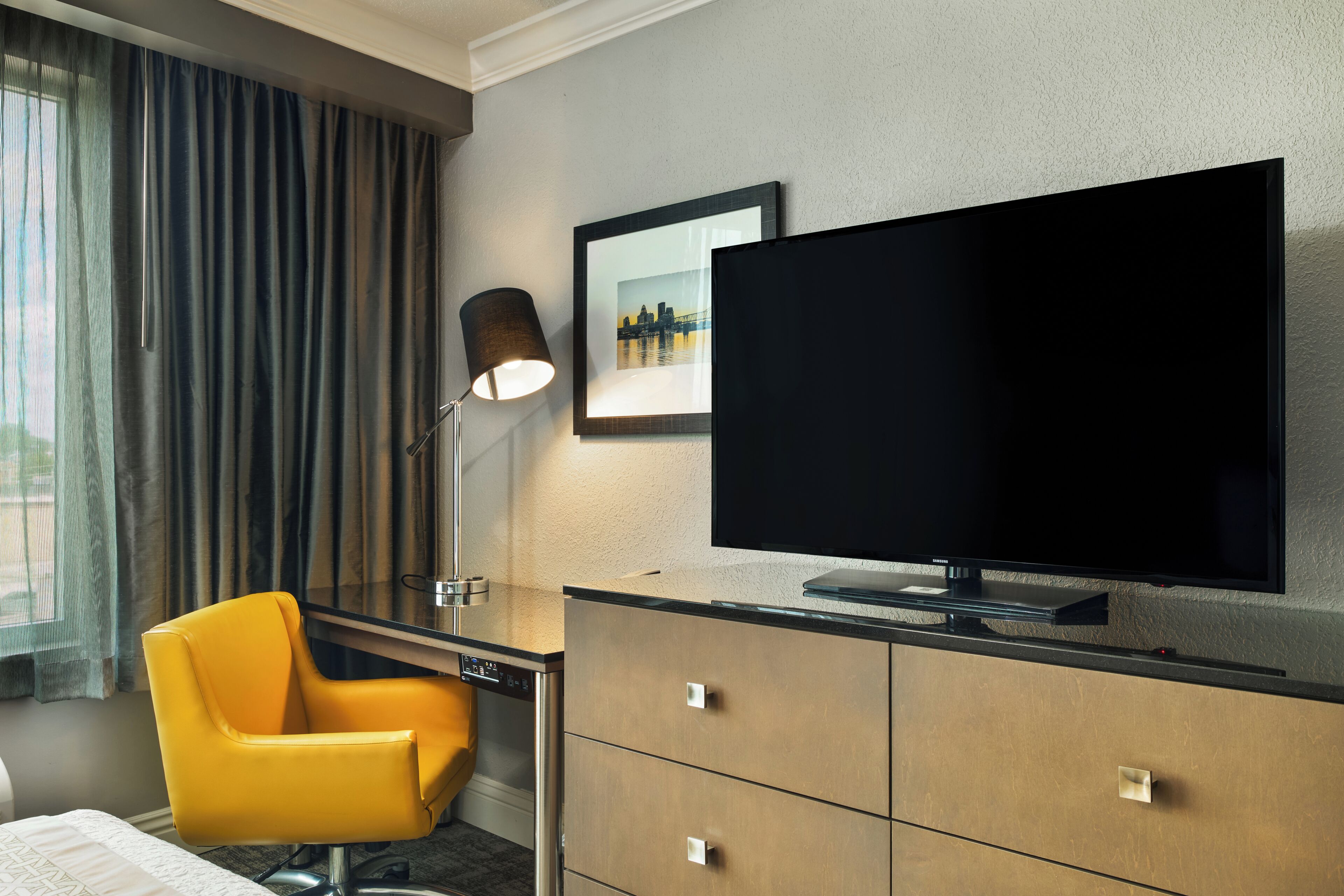 A hotel room with a bed, a desk with a chair, a television, and a framed picture on the wall.