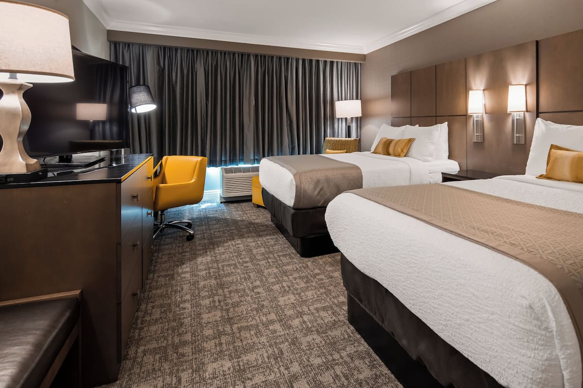 Quarto de hotel com duas camas, uma escrivaninha com computador, uma cadeira e uma janela grande com cortinas.