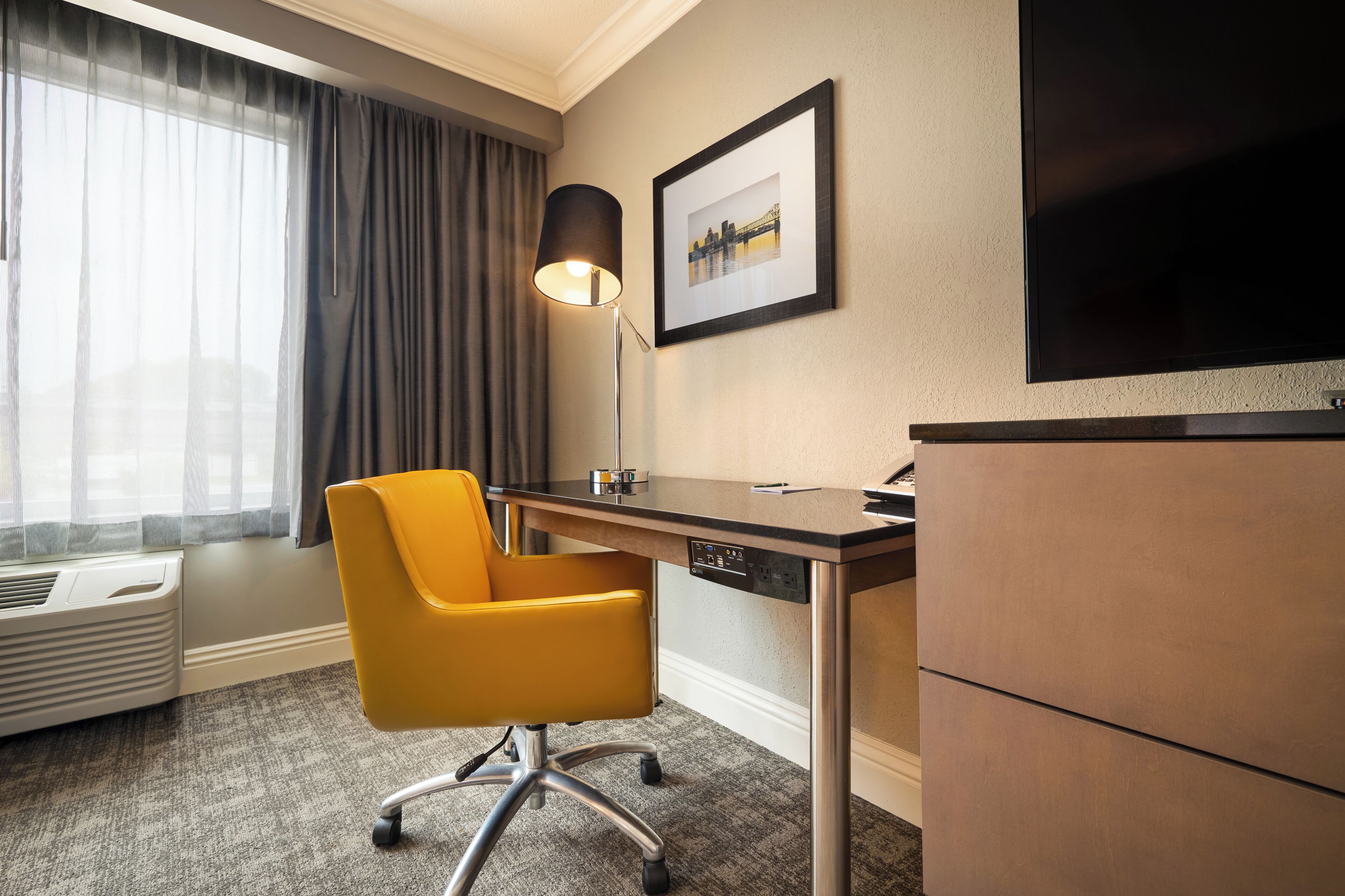 A hotel room with a desk, chair, lamp, and a flat-screen TV.