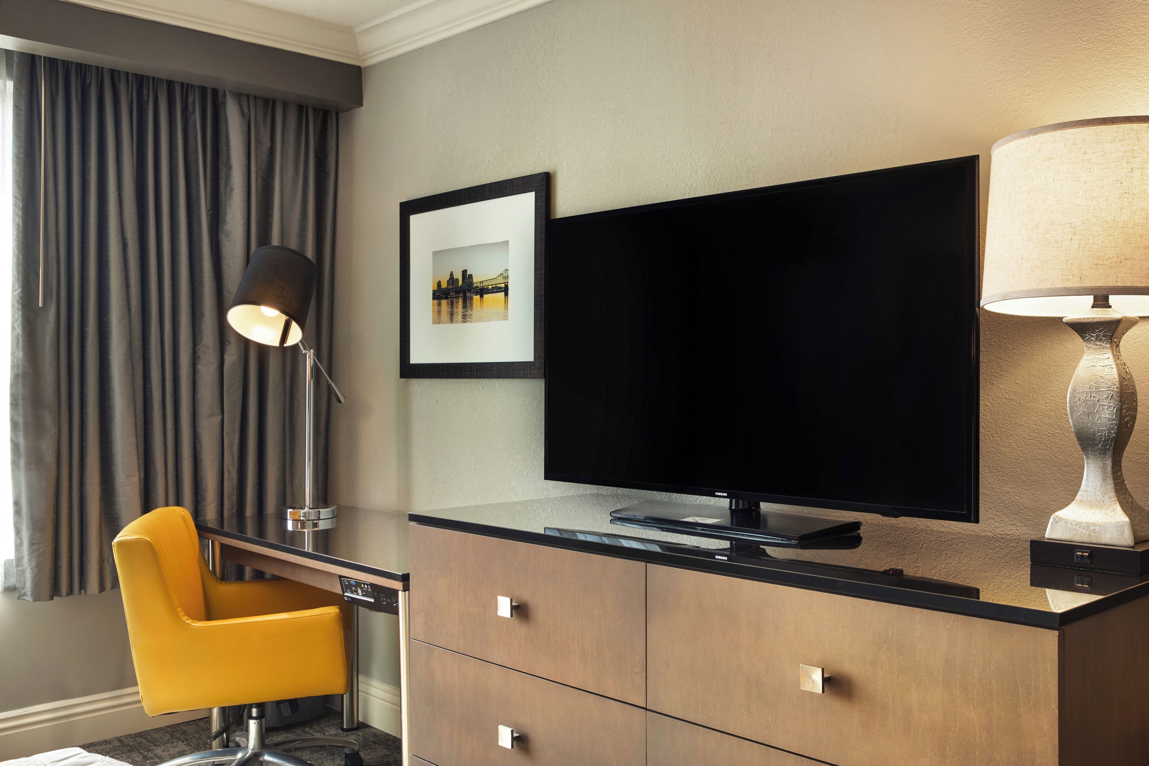 A hotel room with a flat-screen TV, a desk with a chair, a lamp, and a framed picture on the wall.