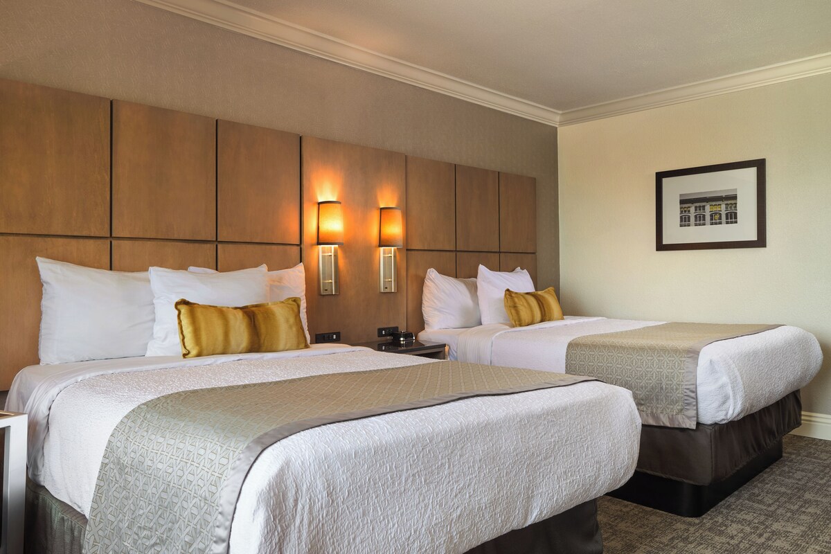 Quarto de hotel com duas camas, cada uma com roupa de cama branca e edredom estampado. Há duas luminárias fixadas na cabeceira e um quadro na parede.