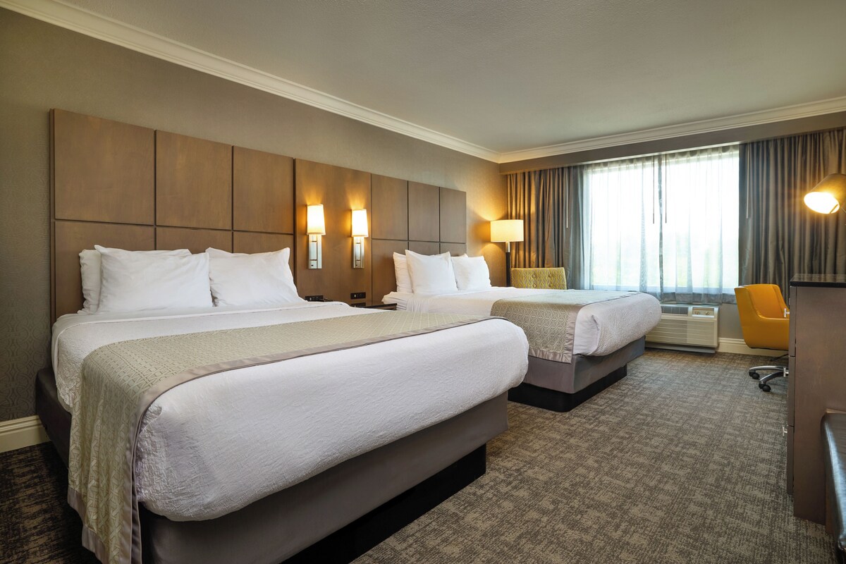Quarto de hotel com duas camas, uma escrivaninha, uma cadeira e uma janela grande com cortinas.