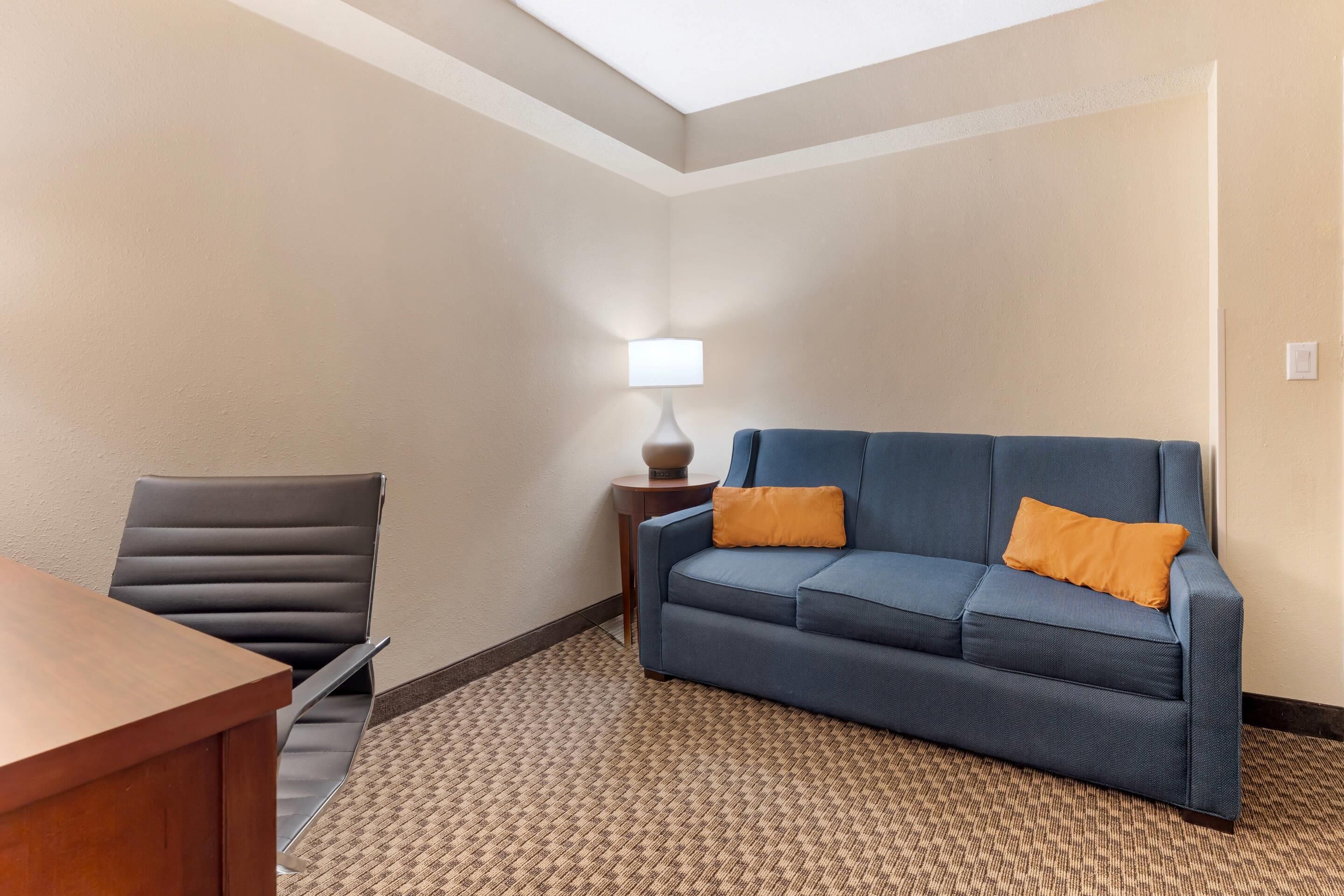 A hotel room with a wooden desk, a blue sofa with orange cushions, a black leather chair, and a patterned carpet.