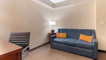 A hotel room with a wooden desk, a blue sofa with orange cushions, a black leather chair, and a patterned carpet.