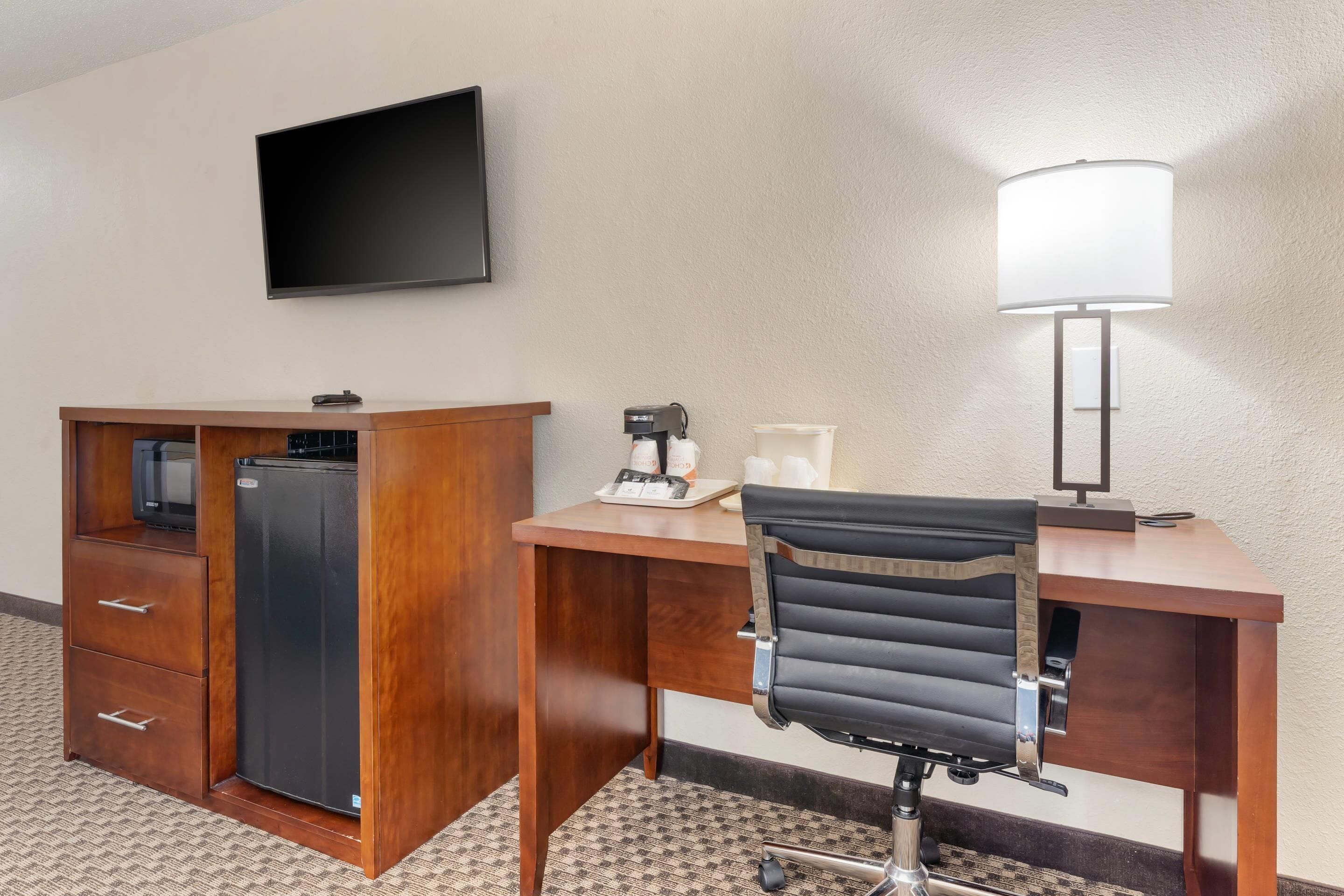 Une chambre d’hôtel équipée d’un bureau en bois, d’un fauteuil de bureau noir, d’un téléviseur à écran plat, d’un réfrigérateur, d’une machine à café et d’une lampe.