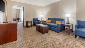 A hotel room with a wooden desk, a blue sofa, two ottomans, a small table, and two lamps. - Comfort Inn Ebensburg (Ebensburg)