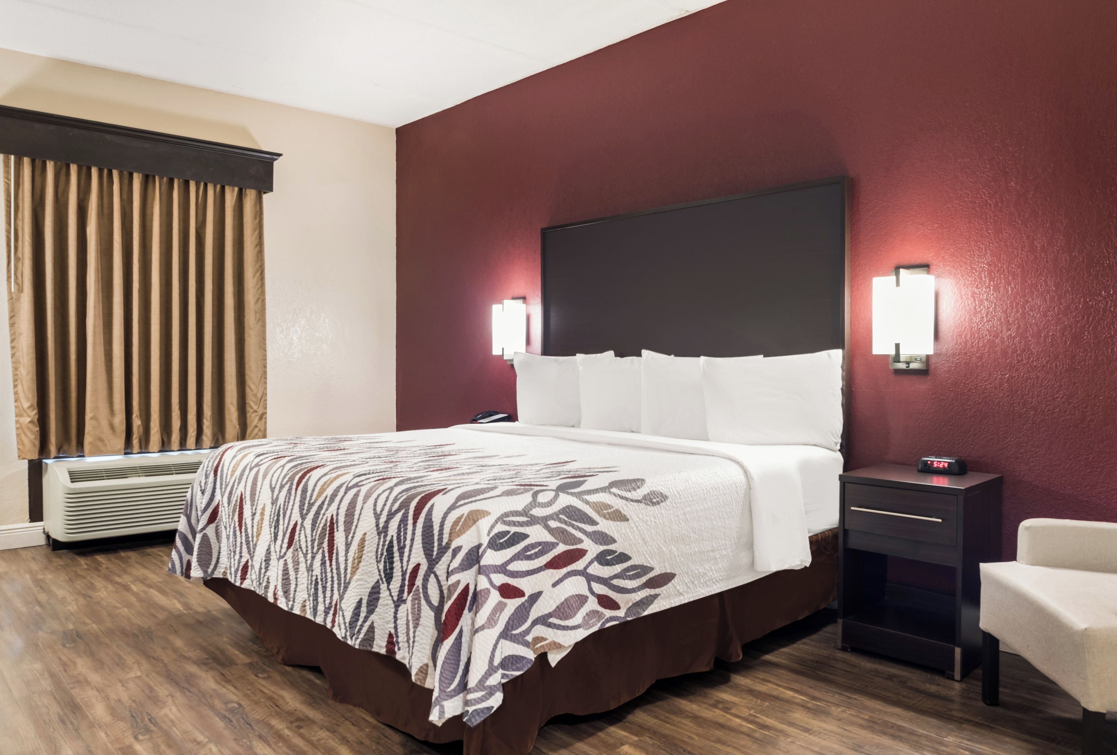 A hotel room with a bed, bedside tables, a nightstand, a chair, a window with curtains, and an air conditioning unit.