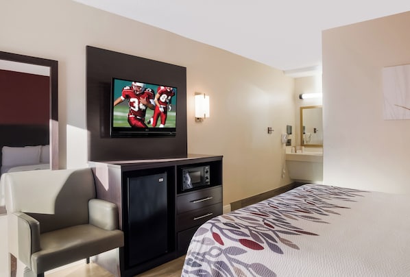 A hotel room with a bed, a TV mounted on the wall, a desk with a chair, a small refrigerator, and a bedside table.