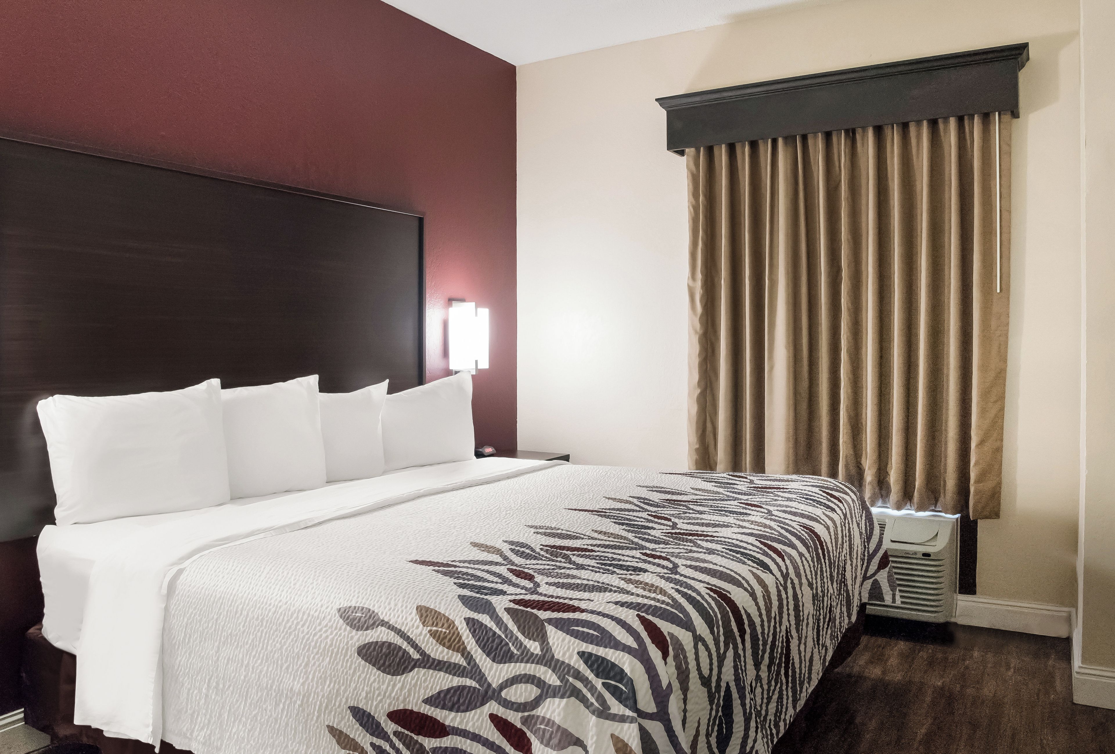 A hotel room with a large bed, a headboard, a window with curtains, and a wall-mounted light.