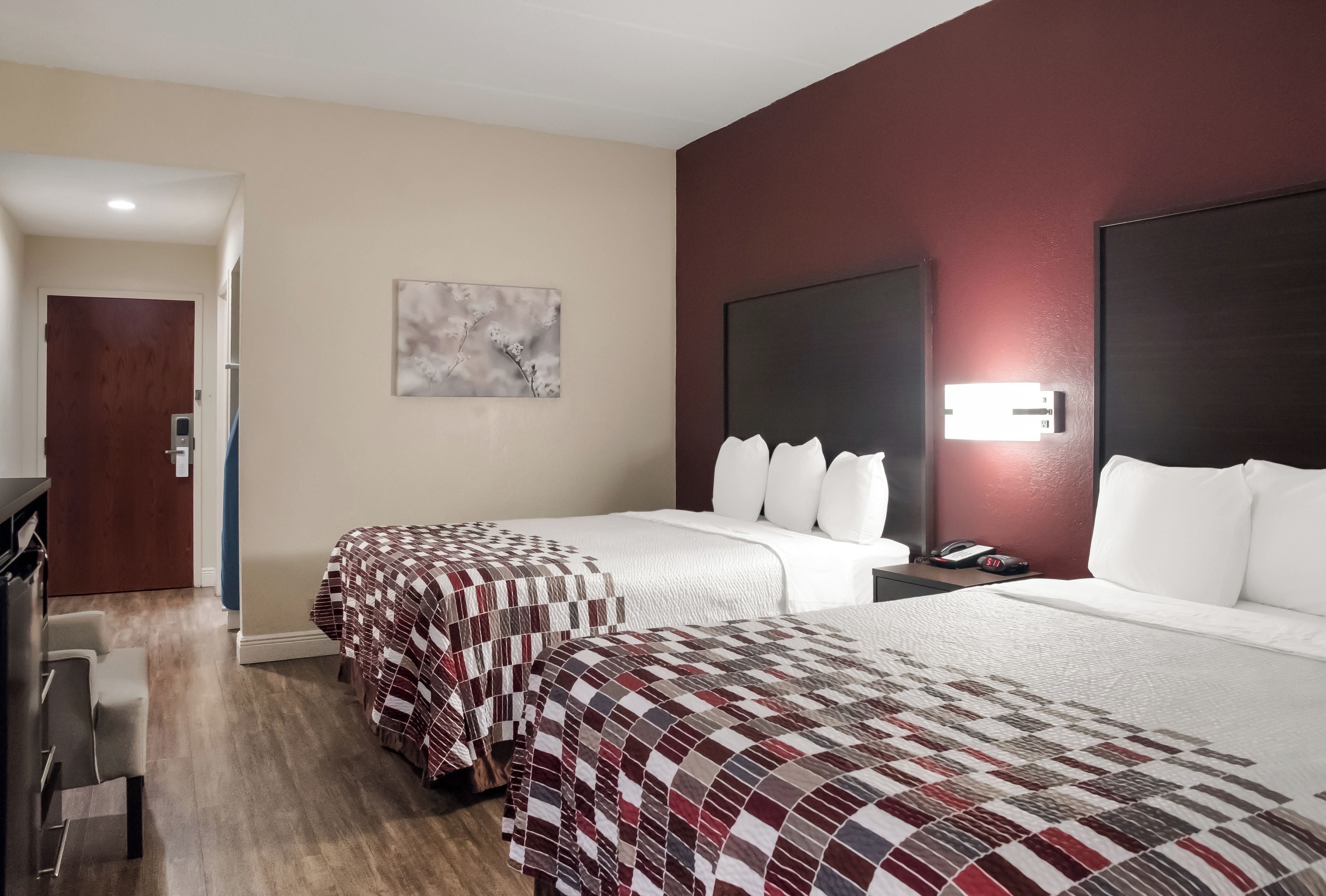 A hotel room with two beds, a desk, a chair, a painting on the wall, and a door leading to another room.