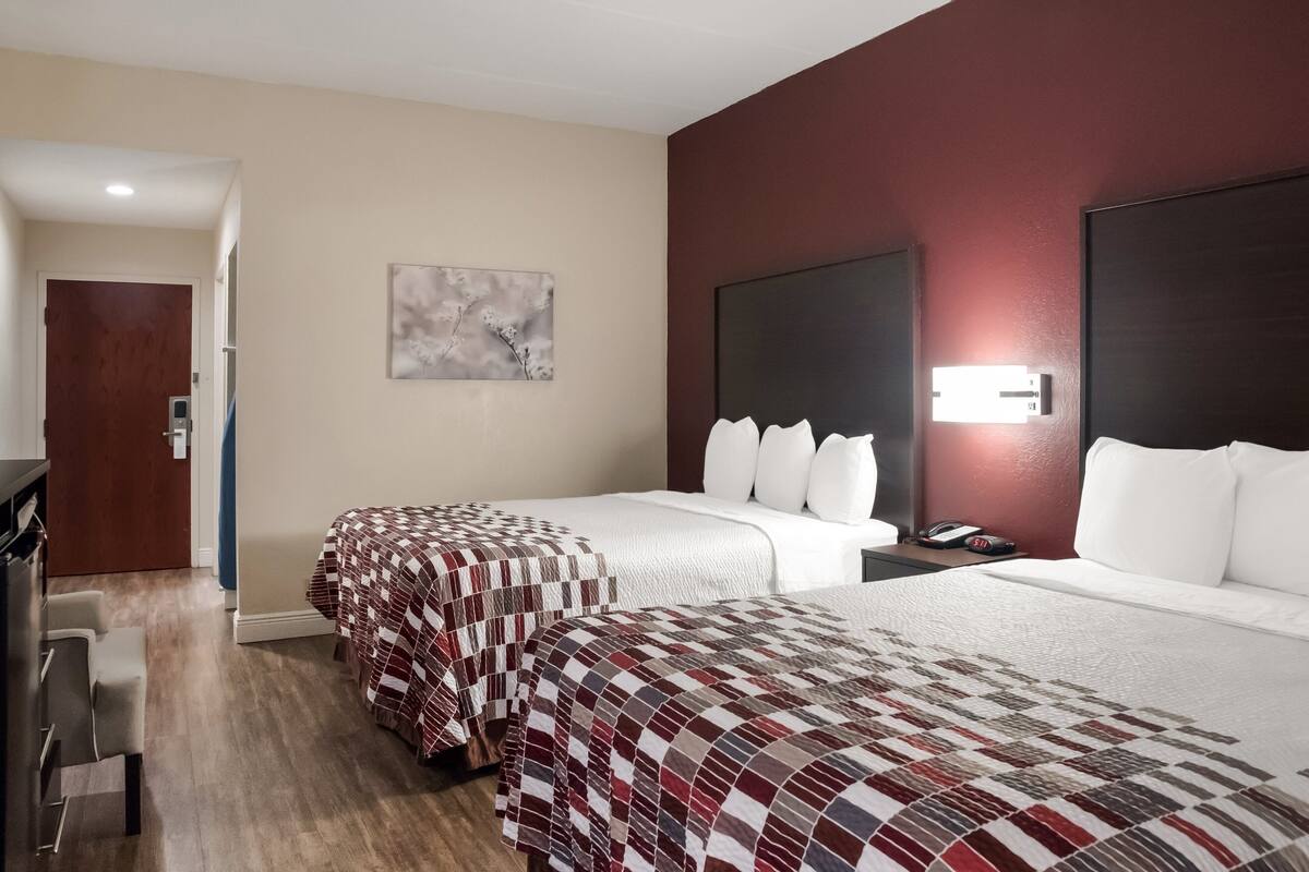 A hotel room with two beds, a desk, and a chair. There is a painting on the wall and a door leading to another room.