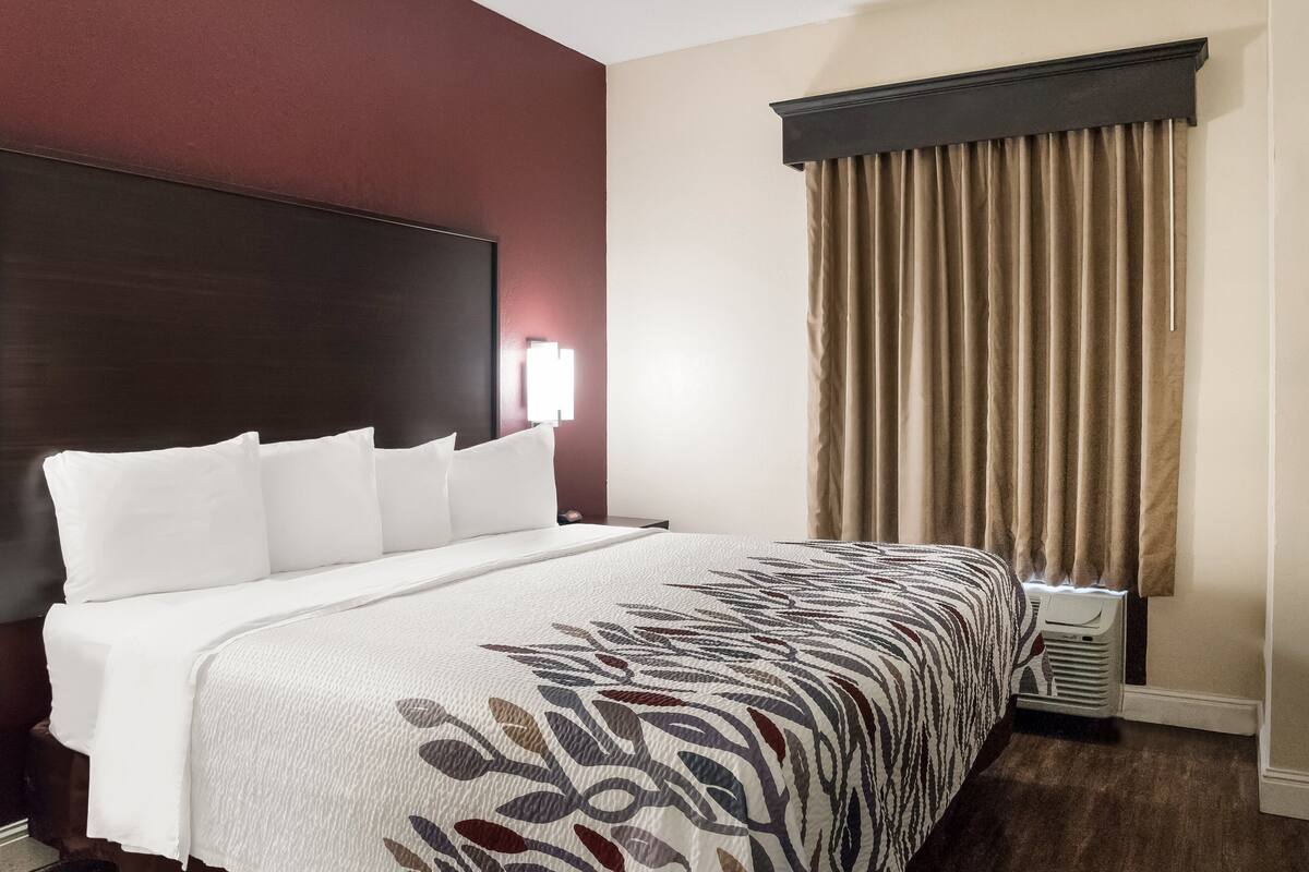 A hotel room with a large bed, a headboard, a bedside lamp, a window with curtains, and a wall-mounted air conditioner.