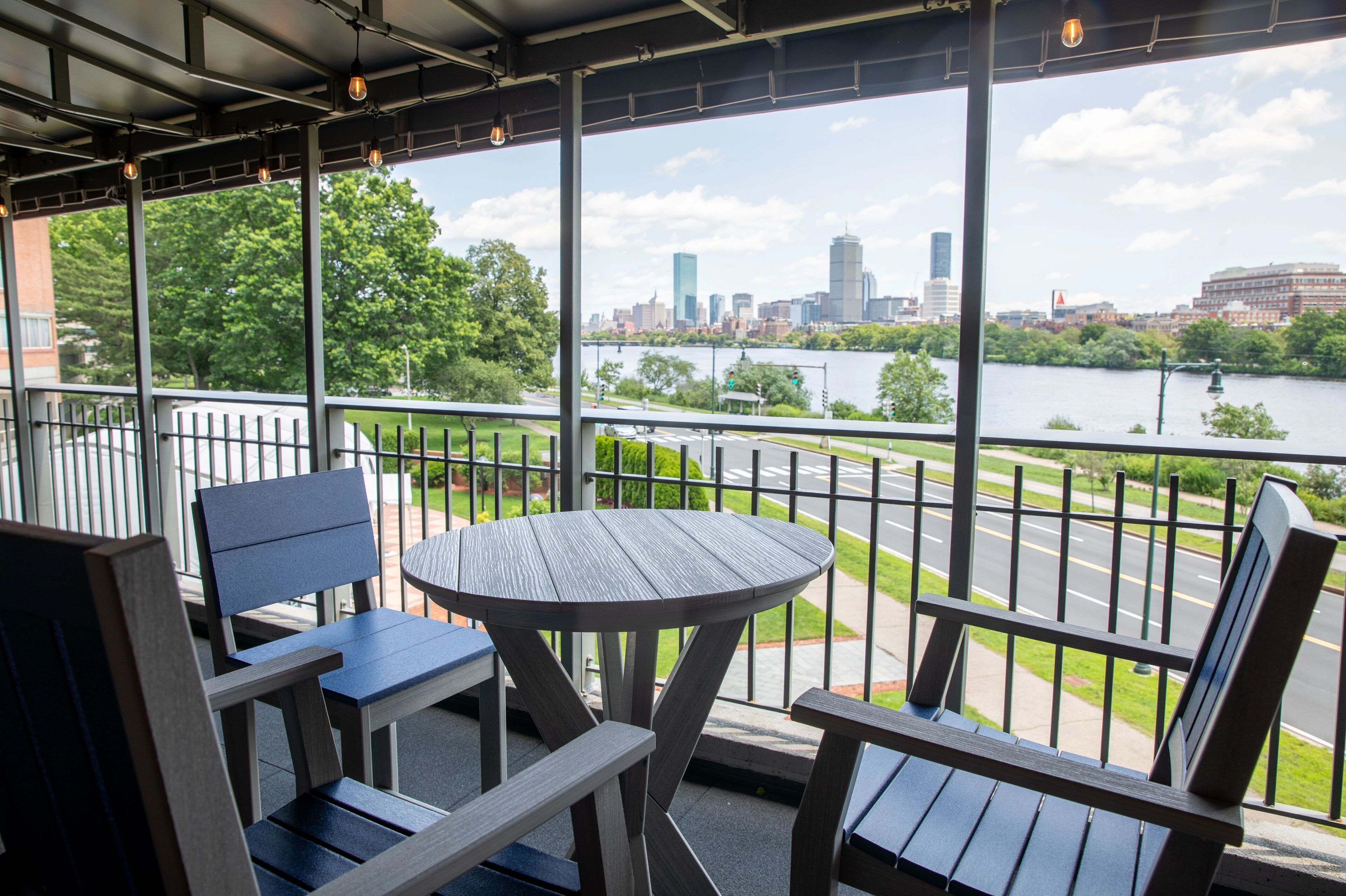 A covered outdoor seating area with a table and chairs, overlooking a river and city skyline.