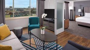 Quarto de hotel moderno com uma grande janela com vista para a cidade, um sofá, uma poltrona azul-turquesa, uma pequena mesa com uma planta, uma cama e uma televisão.