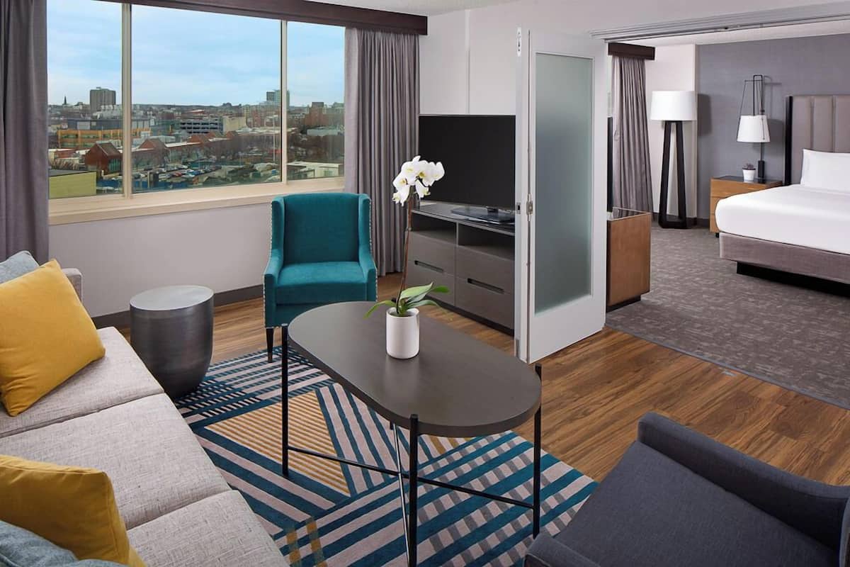 Habitación de hotel moderna con un ventanal grande que ofrece vista a la ciudad, un sofá, un sillón verde azulado, una mesita con una planta, una cama y un televisor.