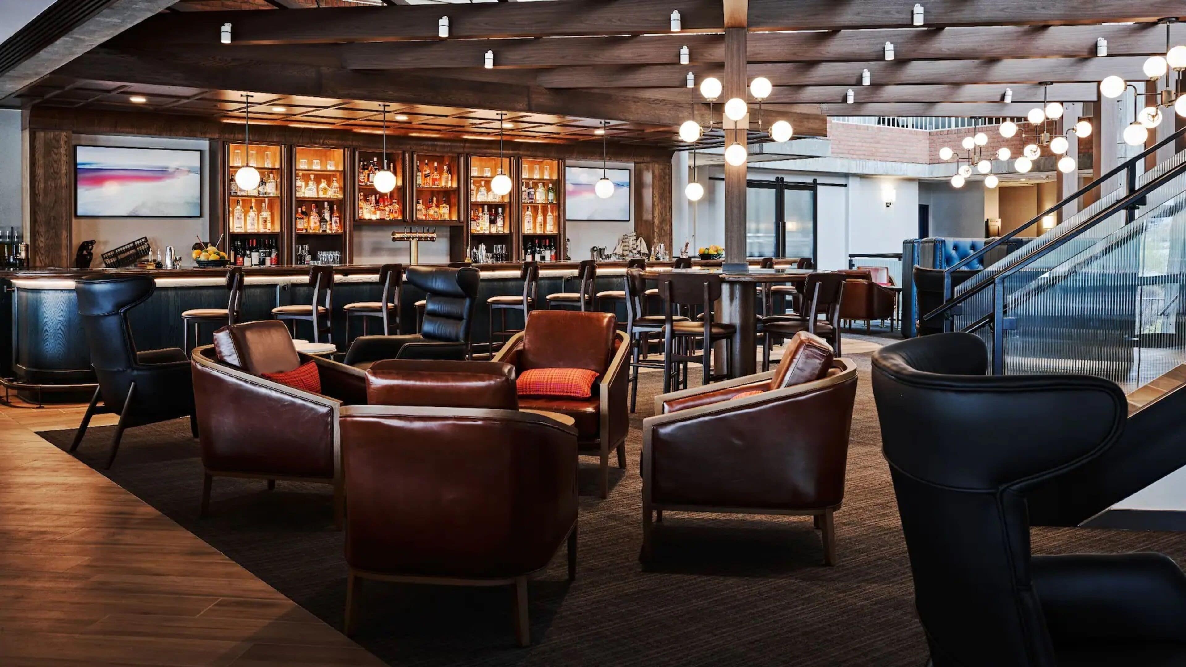 A modern bar area with leather seating, a bar counter with liquor bottles, and a staircase in the background.