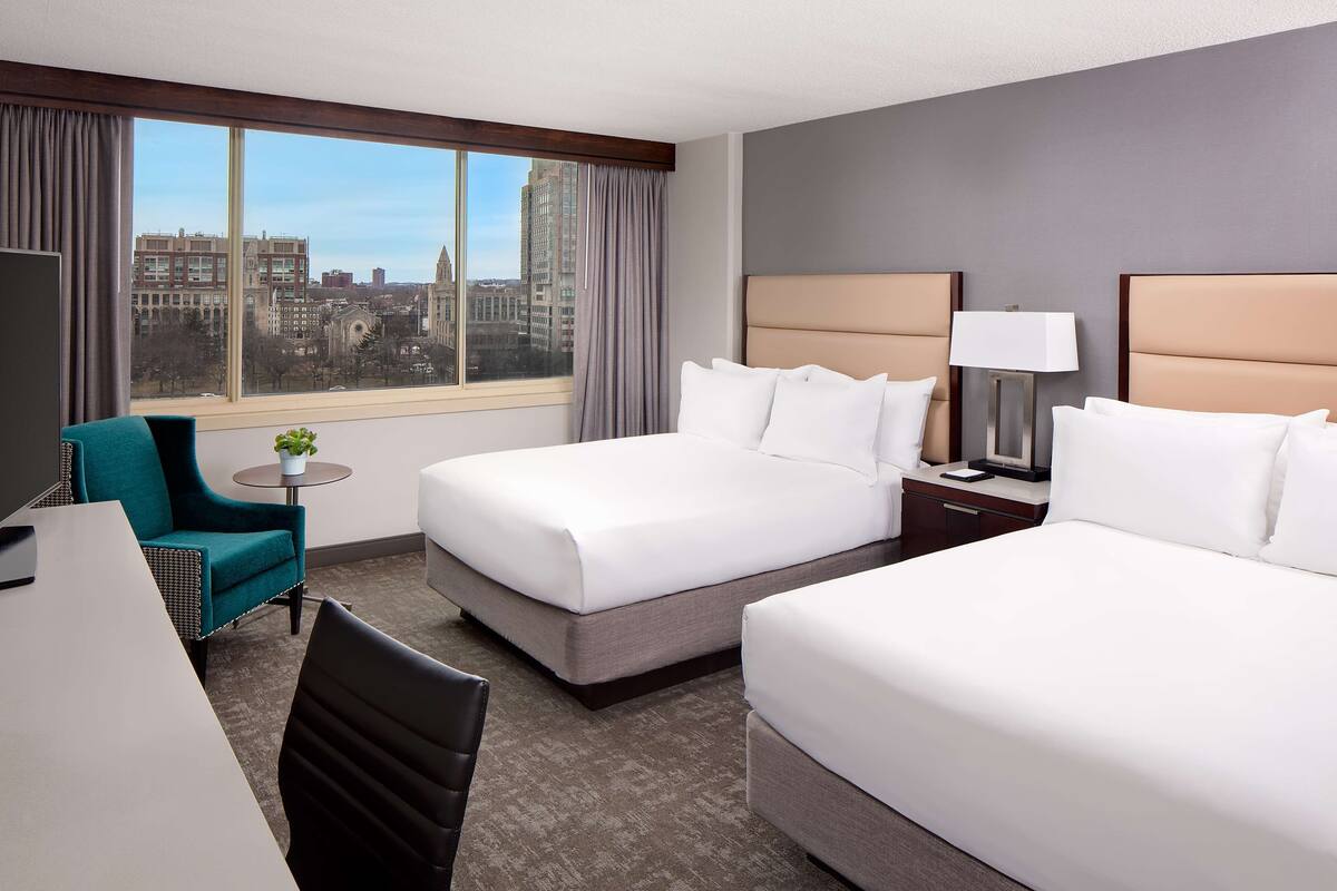 Habitación de hotel con dos camas, un escritorio, una silla, una lámpara y vistas a la ciudad.