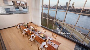 Uma sala de jantar com uma mesa de madeira comprida posta para seis, grandes janelas com vista para a cidade e vista para um rio.