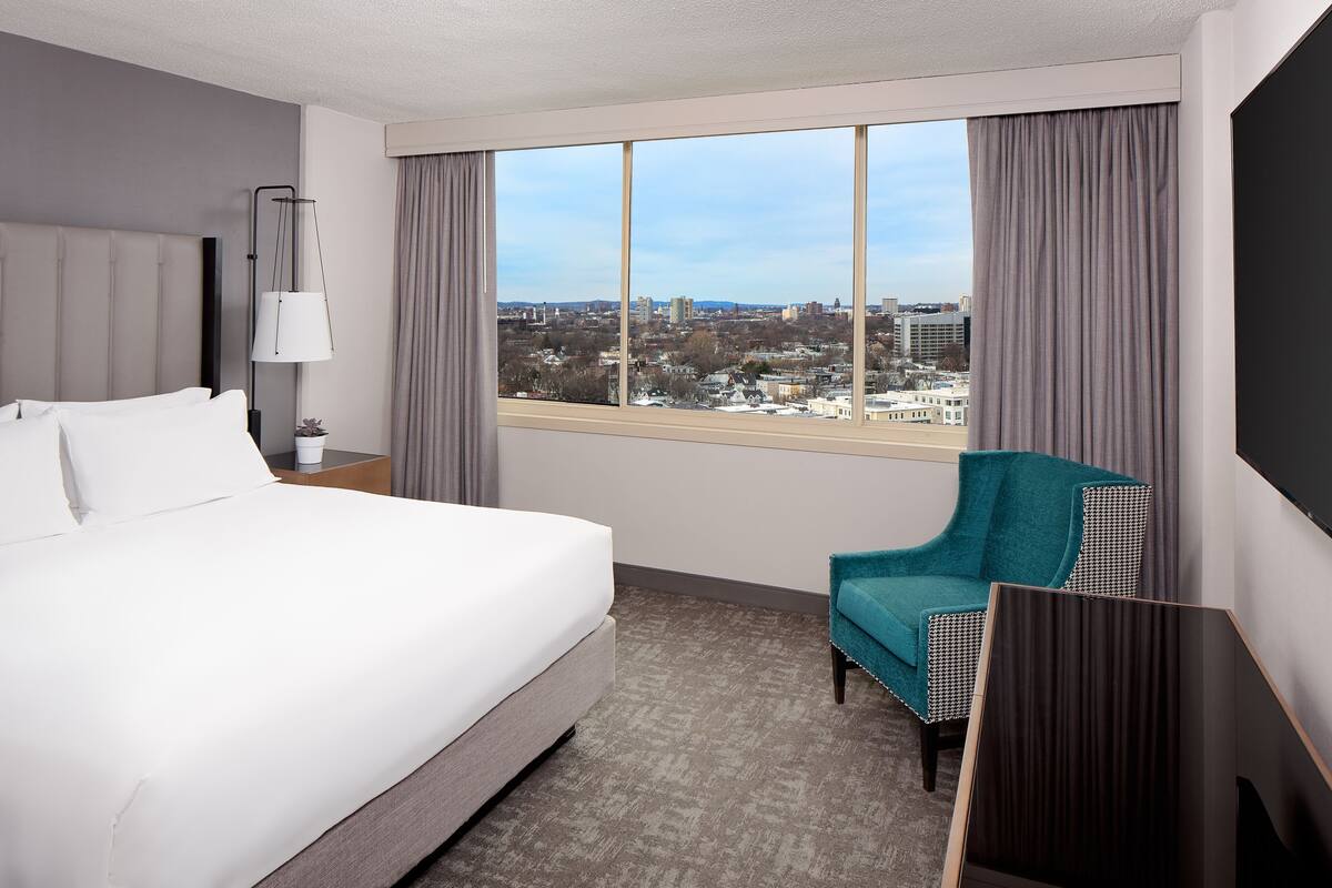 Una habitación de hotel con una cama grande, vistas de la ciudad, una silla color turquesa y una lámpara.
