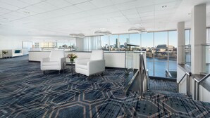 Um amplo hall de hotel com design moderno, com uma grande área de estar, escadas para um andar superior e vista do horizonte da cidade através de amplas janelas.