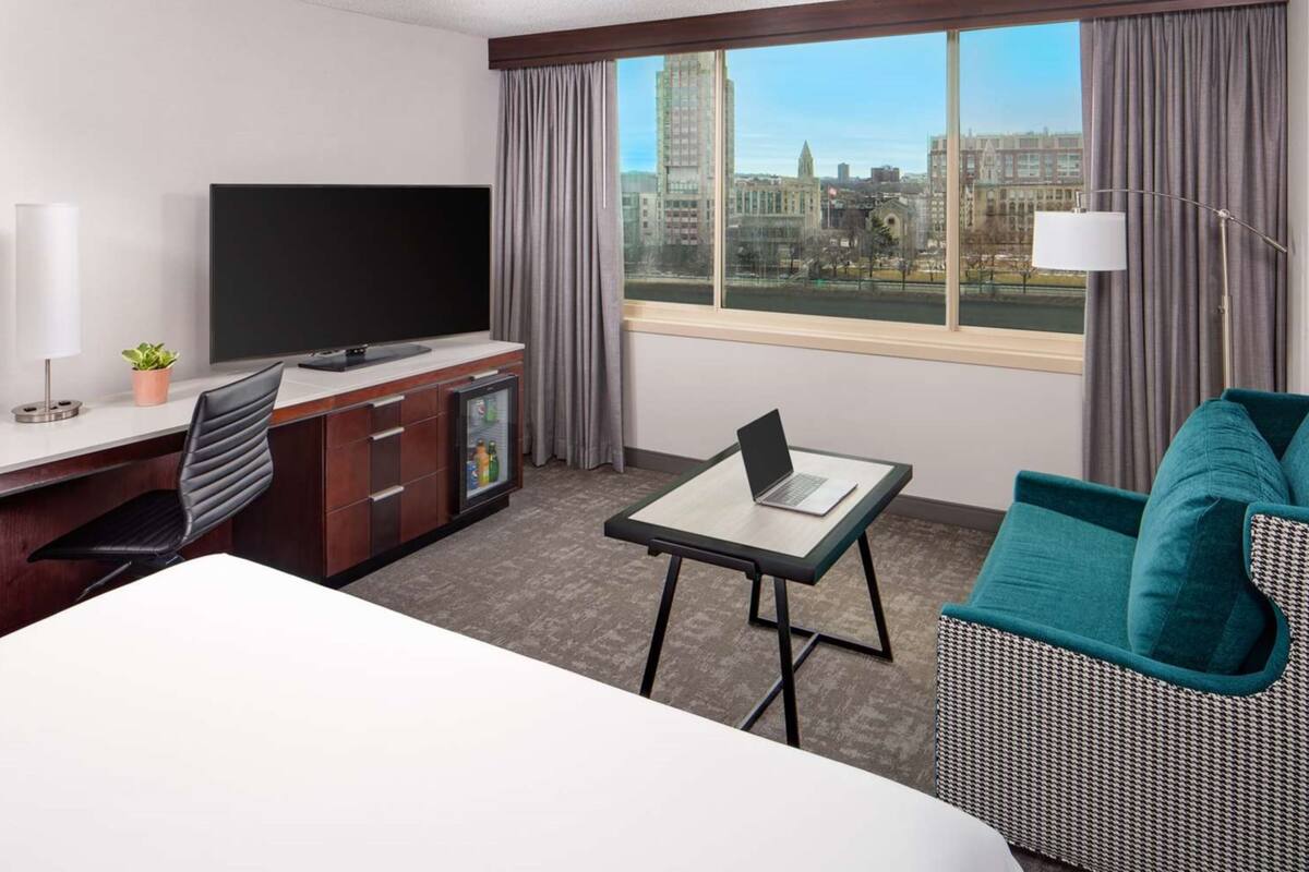Habitación de hotel con un ventanal grande, un televisor de pantalla plana, un escritorio con una computadora portátil, una silla y un sofá.