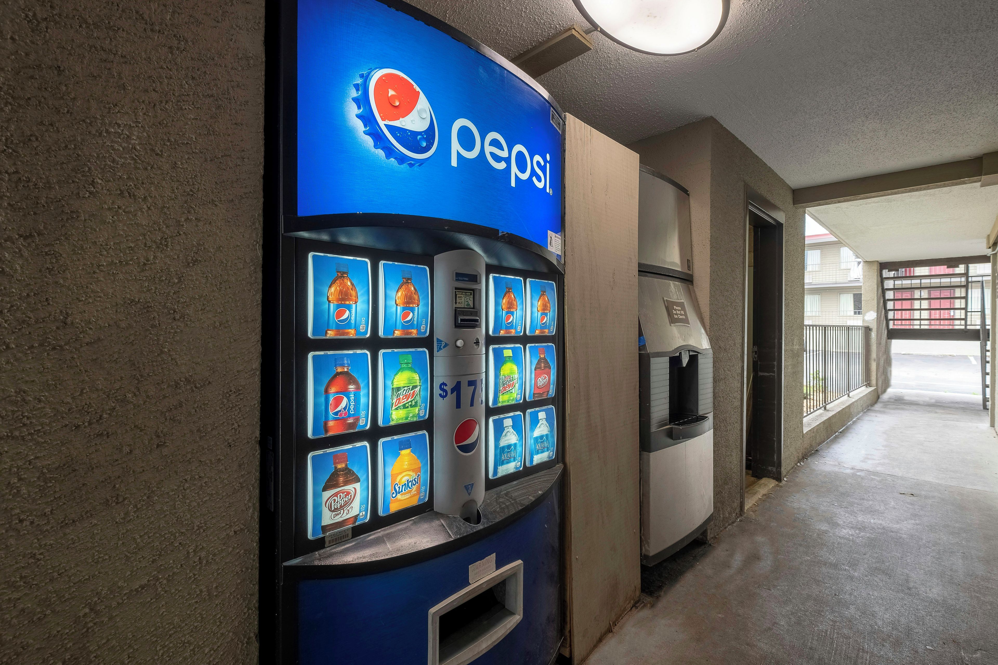 Máquina expendedora con diversas opciones de bebidas, incluyendo Pepsi, en un pasillo con piso de concreto y una puerta que da a otra área.