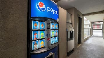 Máquina expendedora con diversas opciones de bebidas, incluyendo Pepsi, en un pasillo con piso de concreto y una puerta que da a otra área.