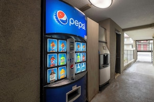 Máquina expendedora con diversas opciones de bebidas, incluyendo Pepsi, en un pasillo con piso de concreto y una puerta que da a otra área.