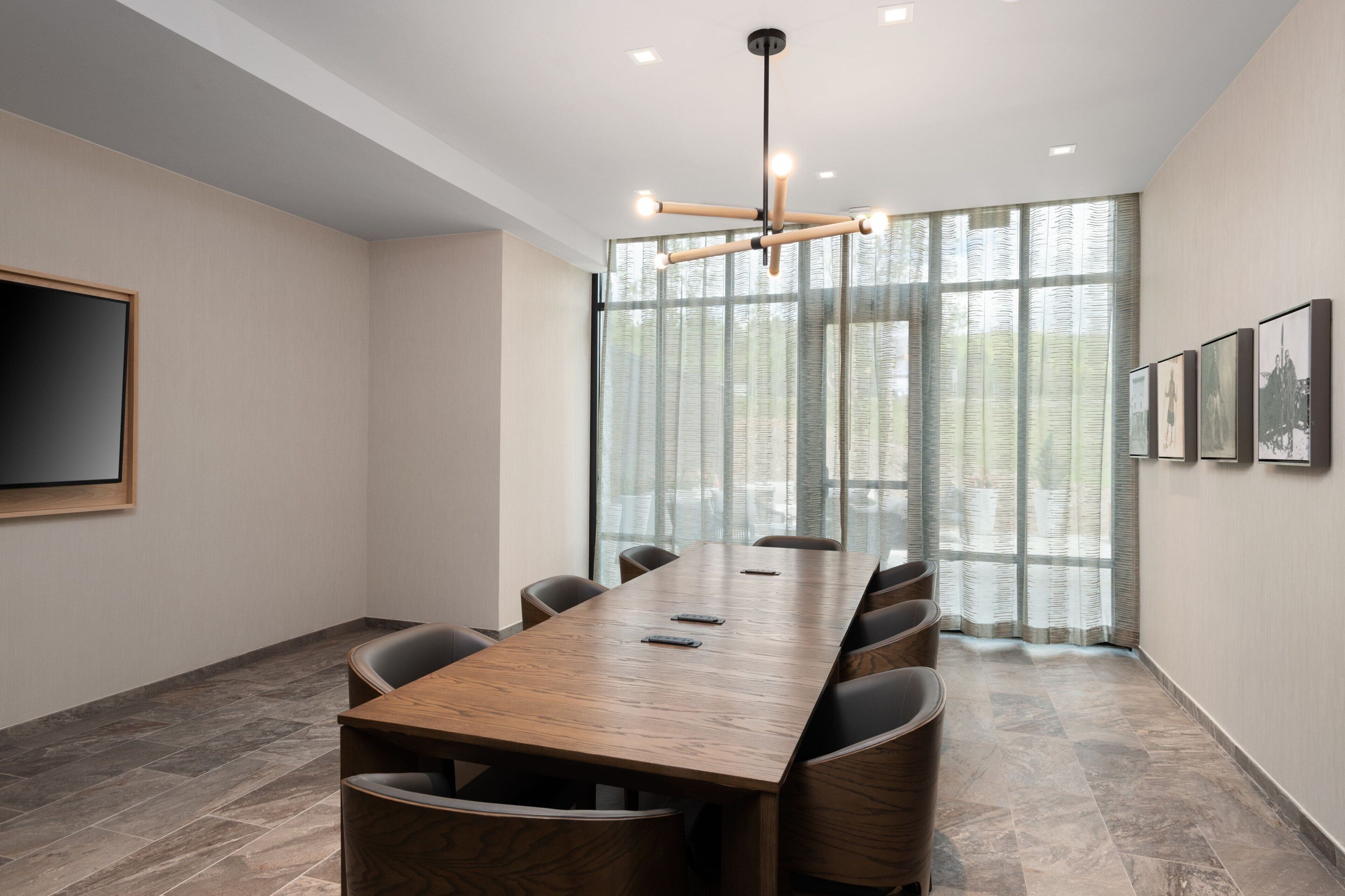 A conference room with a long wooden table, chairs, a flat-screen TV, and large windows with curtains.