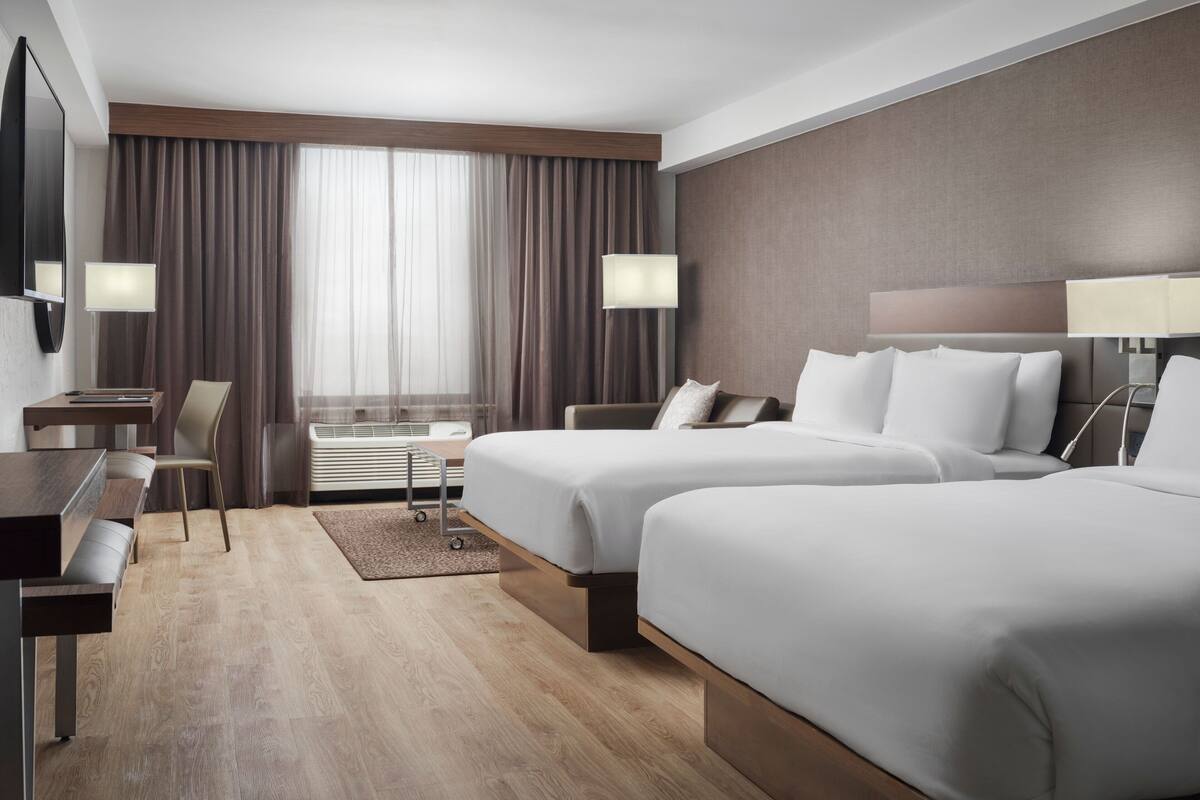 A hotel room with two beds, a desk, a chair, a TV, and a window with curtains.