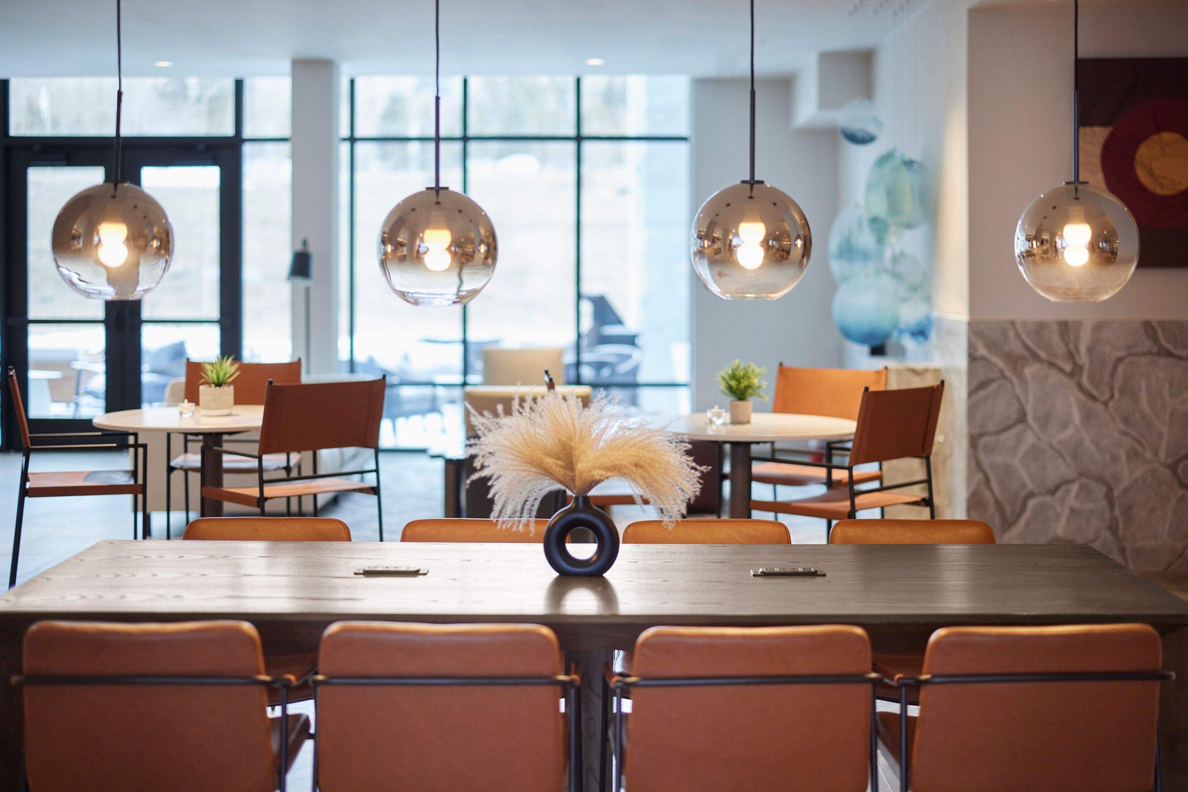 Un comedor moderno con mesas redondas y sillas naranjas, ventanas amplias y lámparas esféricas colgantes.