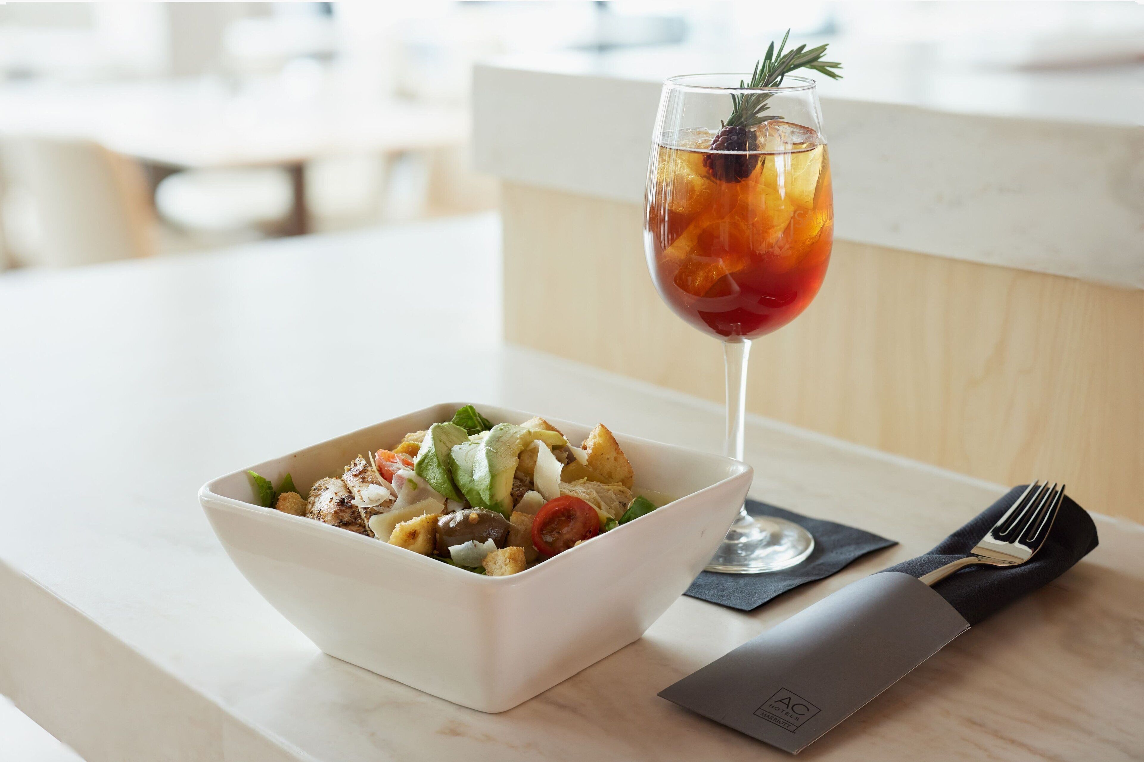 A white bowl of salad with a glass of red drink and a knife on a marble table.