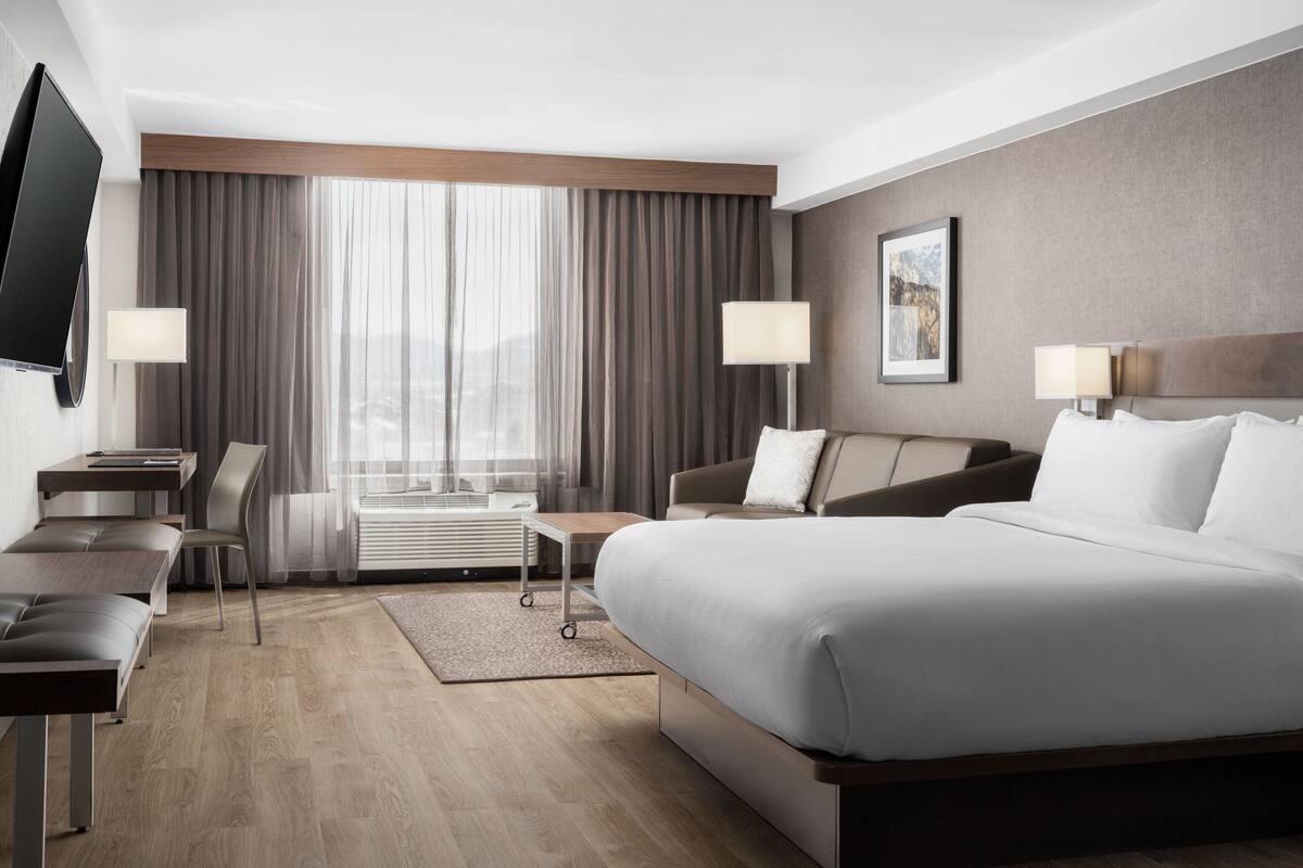 A modern hotel room with a large bed, a desk, a chair, a TV, and a window with curtains.